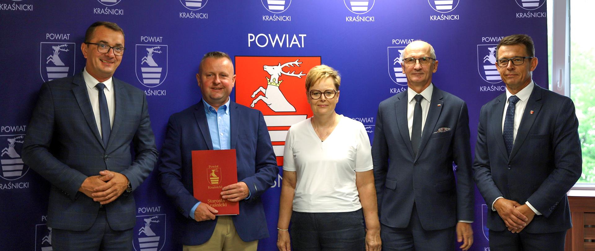 Pamiątkowe zdjęcie z podpisania umowy. Pięć osób stojących w rzędzie obok siebie pozuje do zdjęcia na tle niebieskiej ścianki reklamowej Powiatu Kraśnickiego ozdobionej wizerunkiem herbu powiatu.