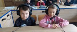 Chłopczyk i dziewczyna siedzą za biurkiem, mają na uszach słuchawki