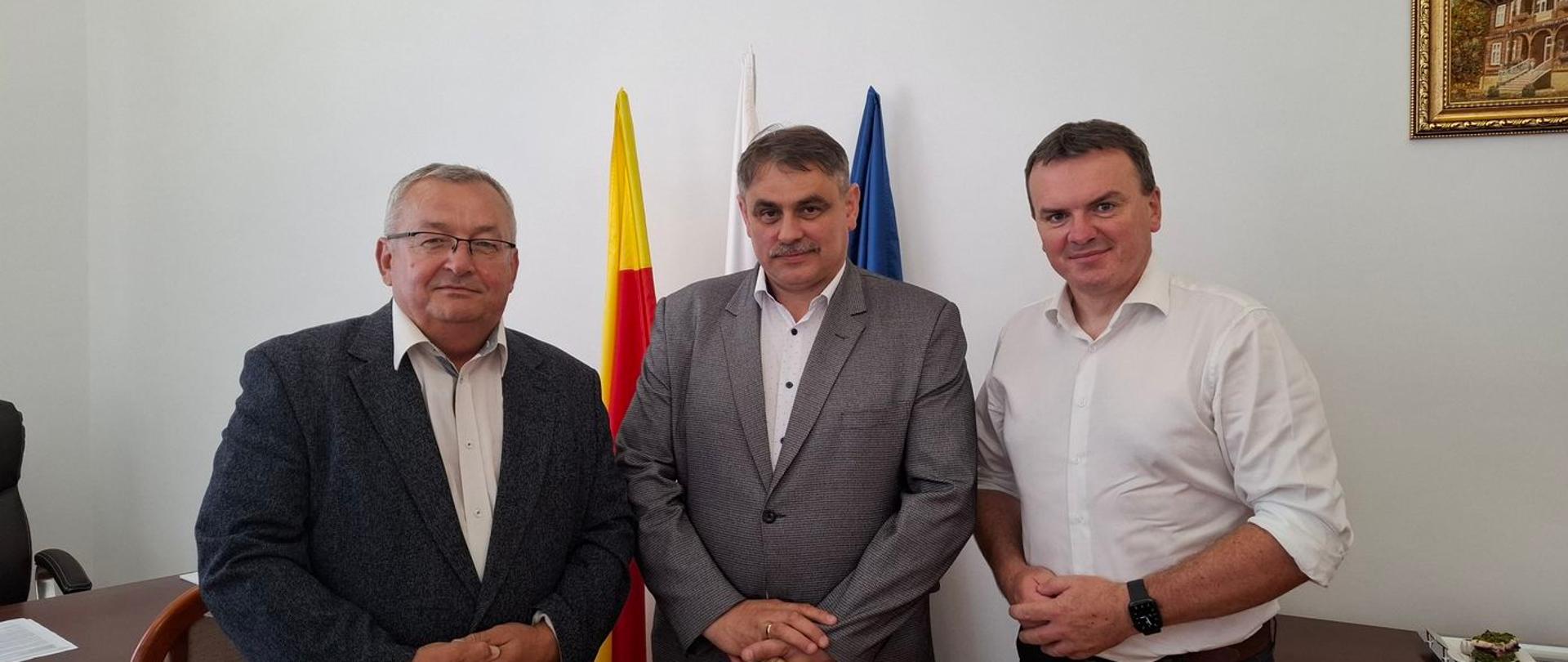 Trzech mężczyzn pozuje na stojąco do zdjęcia, za nimi biała ściana z godłem oraz flagi.