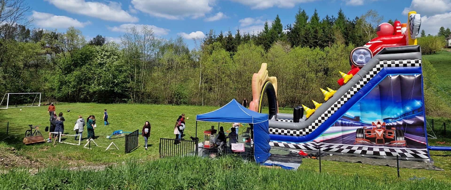 Na zielonym boisku, w otoczeniu drzew, uczestnicy majówki, duża dmuchana zjeżdżalnia oraz stoisko gastronomiczne. 