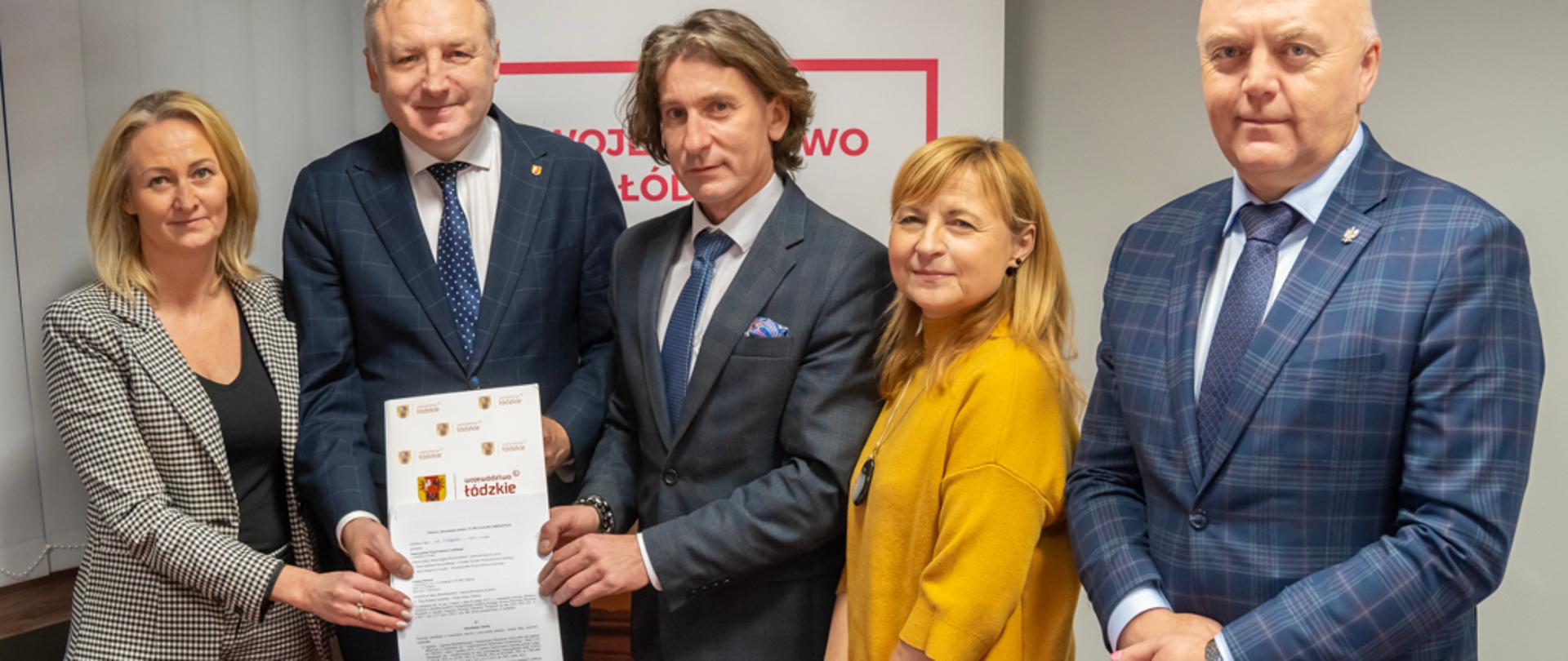 Pięć osób stoi, trzy z nich trzymają zadrukowaną kartkę odwróconą treścią w kierunku obserwatora. W tle baner.