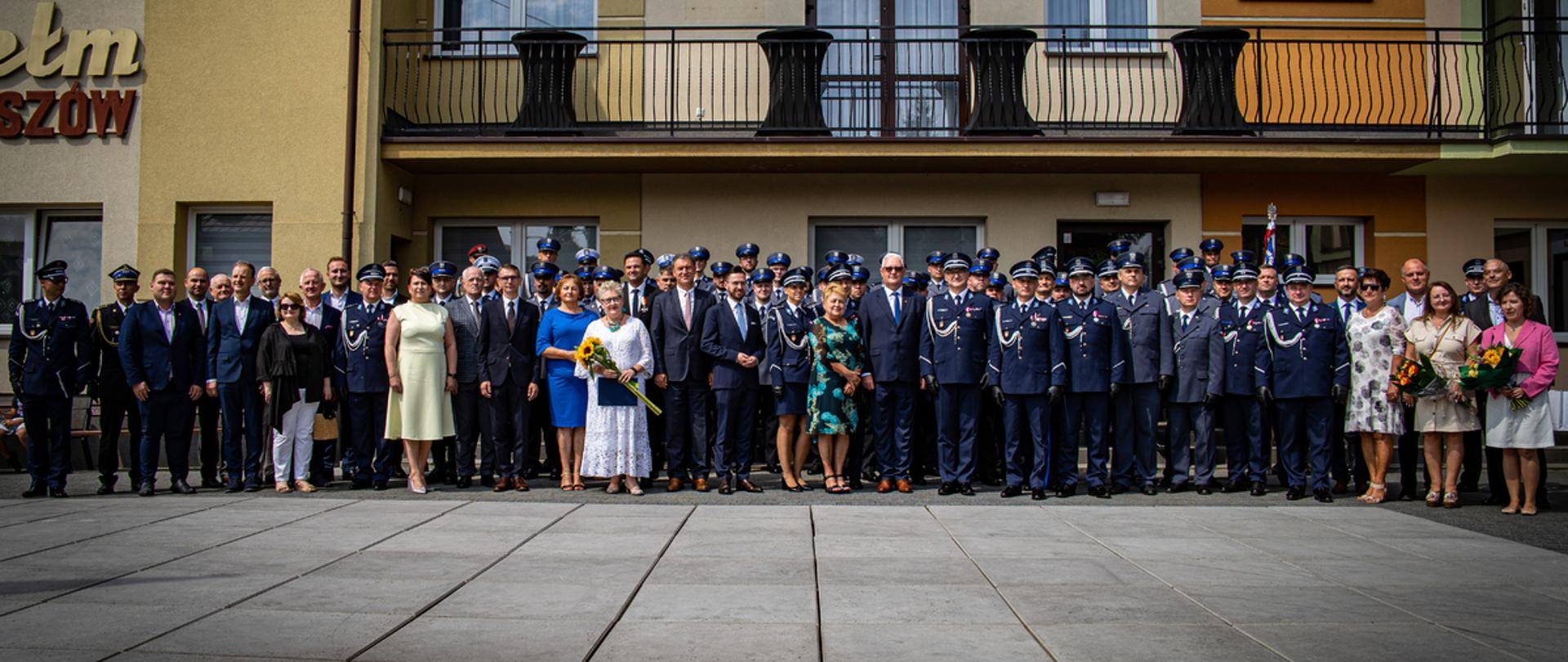 Na tle budynku stoi grupa ludzi elegancko ubranych wśród których znajdują się Policjanci w mundurach. 