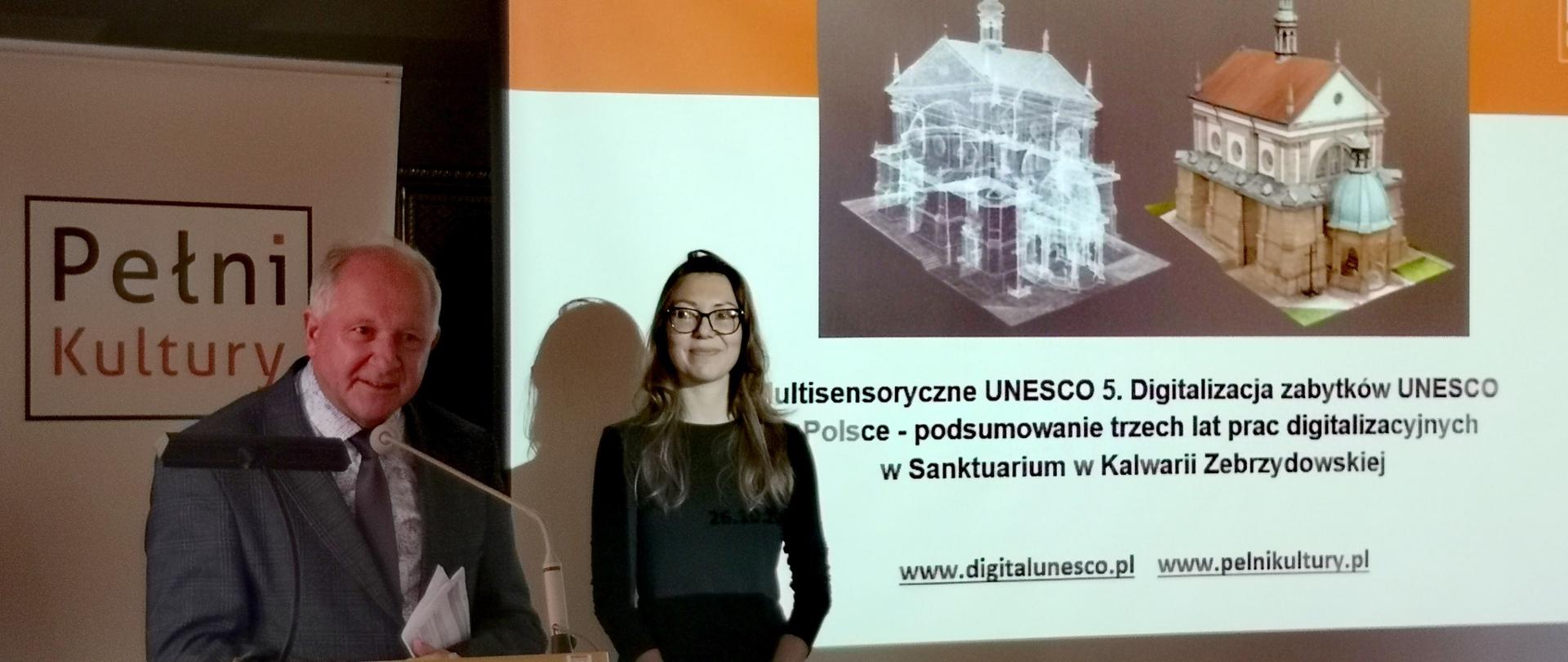 Stojący przy mównicy w zaciemnionej sali burmistrz wypowiada się do mikrofonu; za nim ubrana na czarno kobieta i wyświetlony na ekranie slajd z prezentacji, przedstawiający trójwymiarowy model kaplicy.