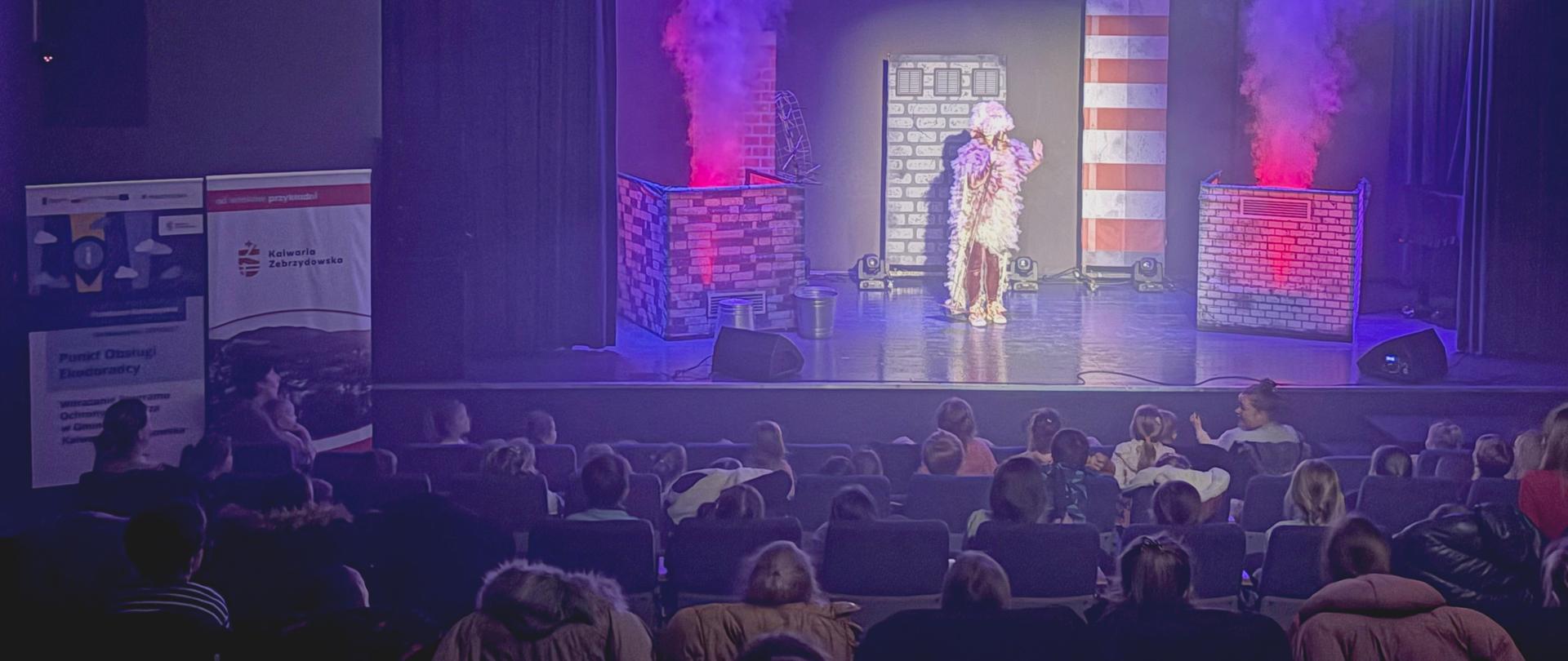 Zgromadzone na sali widowiskowej dzieci oglądają w półmroku przedstawienie; na scenie aktorka w ptasim stroju i kominy, z których wydobywa się kolorowy dym
