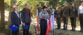 Uroczystość przy pomniku w lesie z udziałem pocztów sztandarowych i uczestników wydarzenia.
