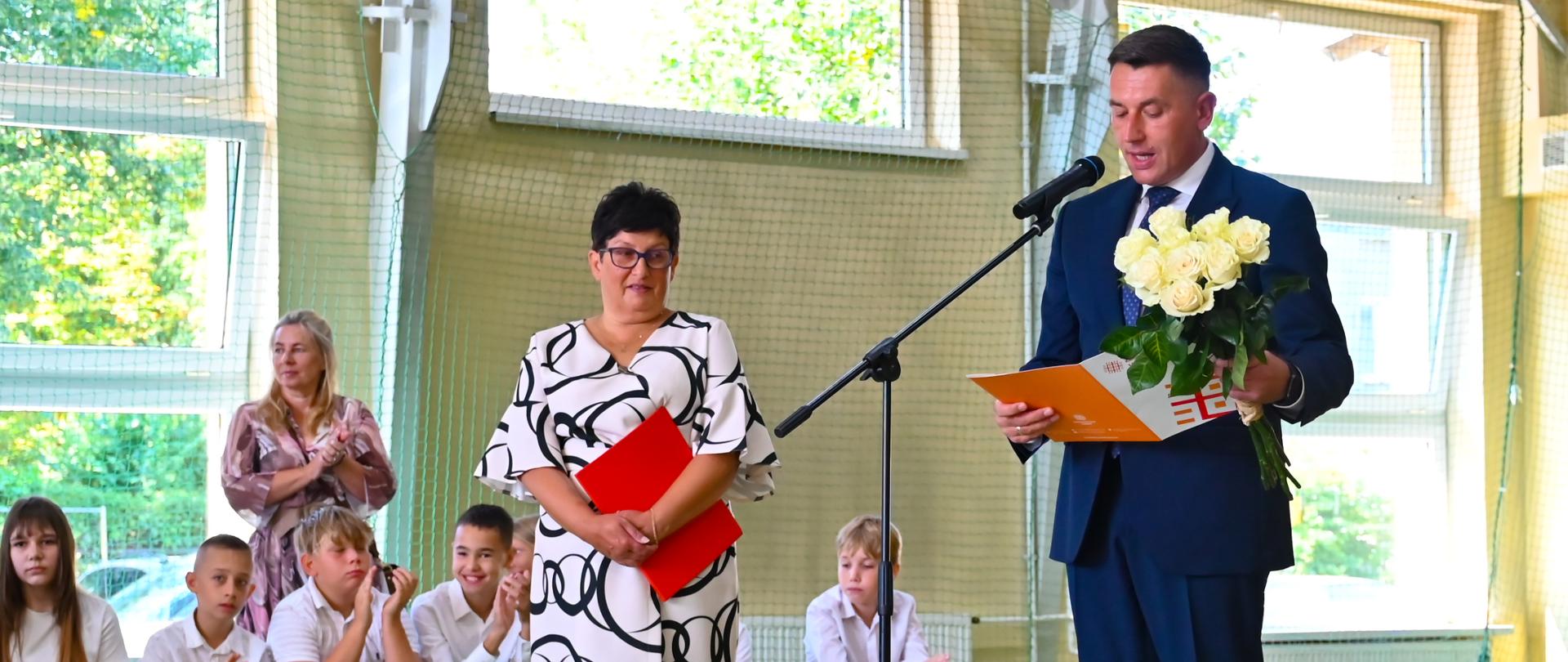 Burmistrz przemawia w szkole, trzyma bukiet białych róż, w tle dyrektorka szkoły i dzieci.