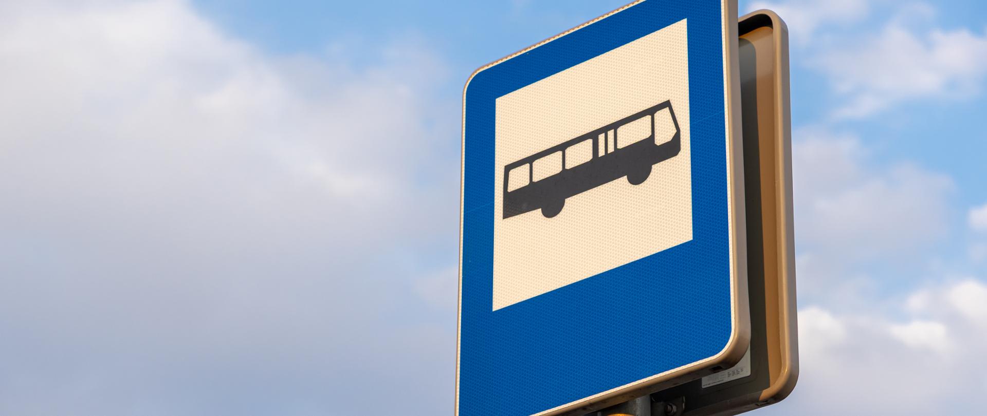 Prostokątny, niebieski znak drogowy D-15 oznaczający przystanek autobusowy z czarnym piktogramem autobusu na białym tle, przedstawiony z dołu na tle lekko zachmurzonego nieba.