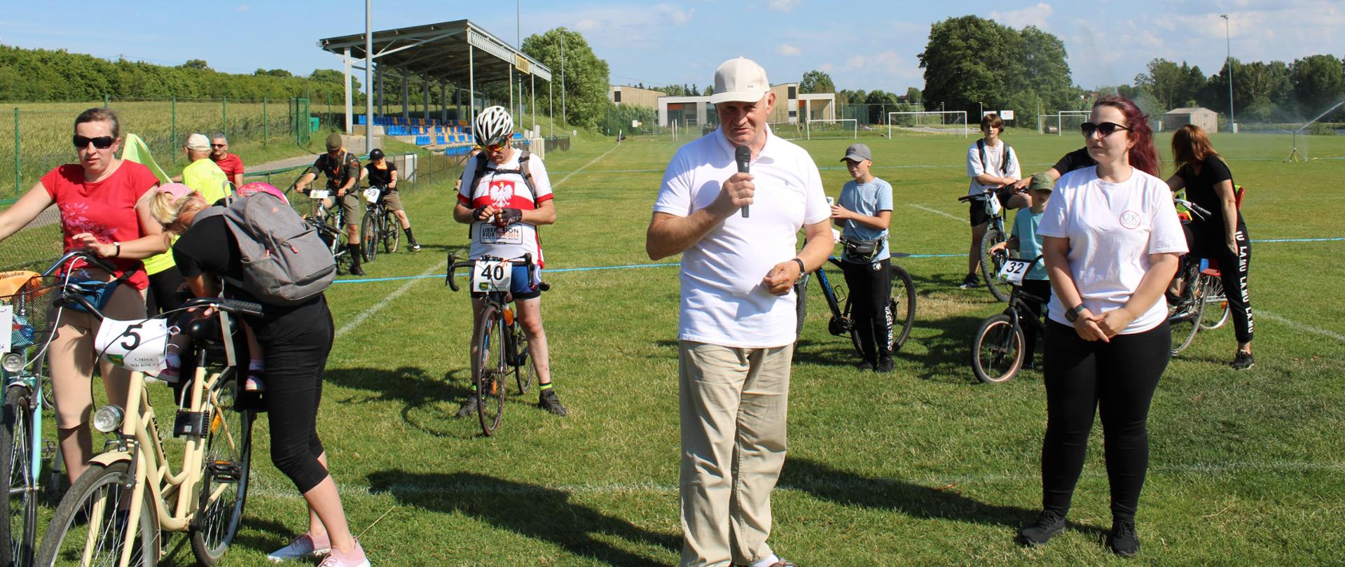 Uczestnicy rajdu stojący z rowerami na murawie stadionu sportowego w Wilkołazie. Pośrodku zdjęcia w białej koszulce, beżowych spodniach i beżowym kaszkiecie na głowie oraz z mikrofonem w ręku stoi Członek Zarządu Powiatu w Kraśniku Eugeniusz Pelak.