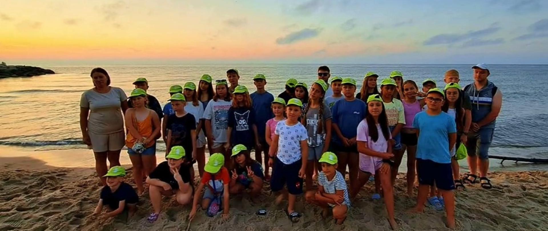 Na zdjęciu widać grupę około 35 osób – głównie dzieci i młodzieży – stojących na plaży o zachodzie słońca. Wszyscy uczestnicy mają na głowach jaskrawe, żółto-zielone czapki z daszkiem, co sugeruje, że należą do jednej grupy zorganizowanej, np. kolonii lub obozu. W tle rozpościera się spokojne morze, a niebo jest w pastelowych odcieniach pomarańczu, różu i błękitu. Po lewej stronie widać fragment skalistego wybrzeża. W górnych rogach zdjęcia znajdują się logotypy – po lewej czerwony znak graficzny, po prawej zielone logo z napisem „Stowarzyszenie Mieszkańców Dolany na Ludowo i Sportowo”.