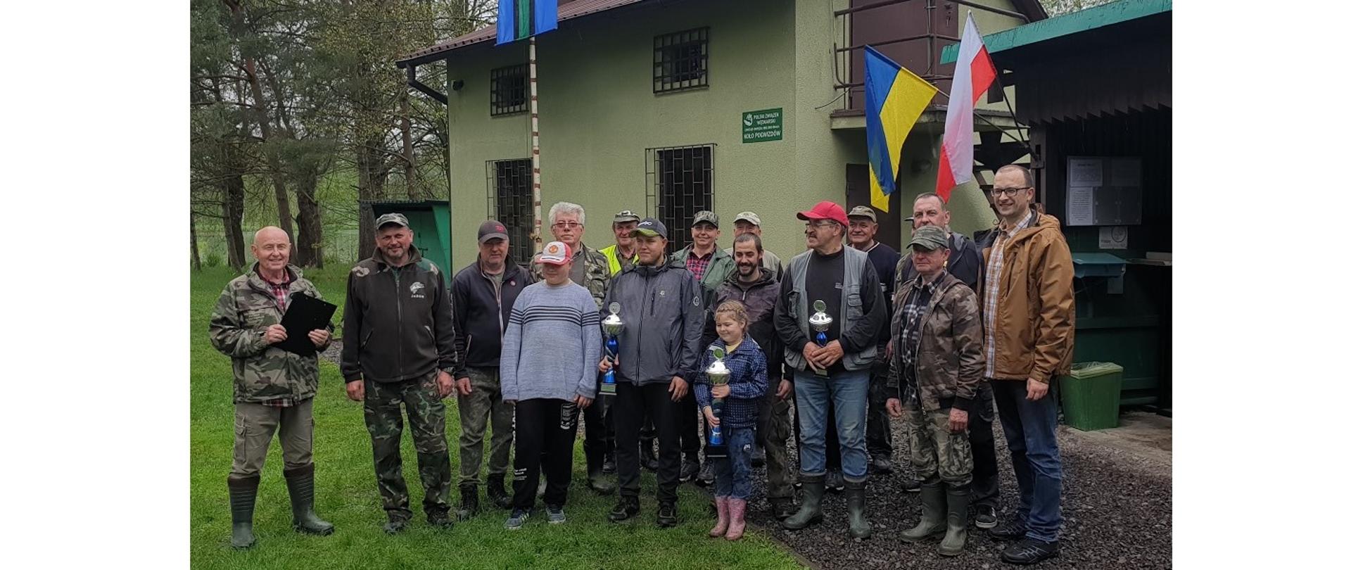 Finaliści Charytatywnych Zawodów Wędkarskich z ich organizatorami. Kilkanaście osób pozuje do zdjęcia. Z tyłu na budynku wiszą dwie flagi narodowe: polska i ukraińska