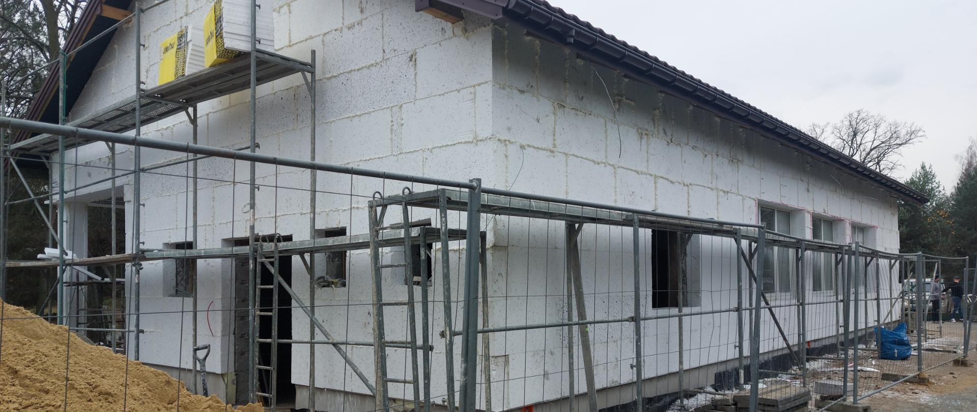 Budynek pokryty białym styropianem. Wokół rozstawione rusztowanie. Budynek ogrodzony tymczasowym ogrodzeniem metalowym.