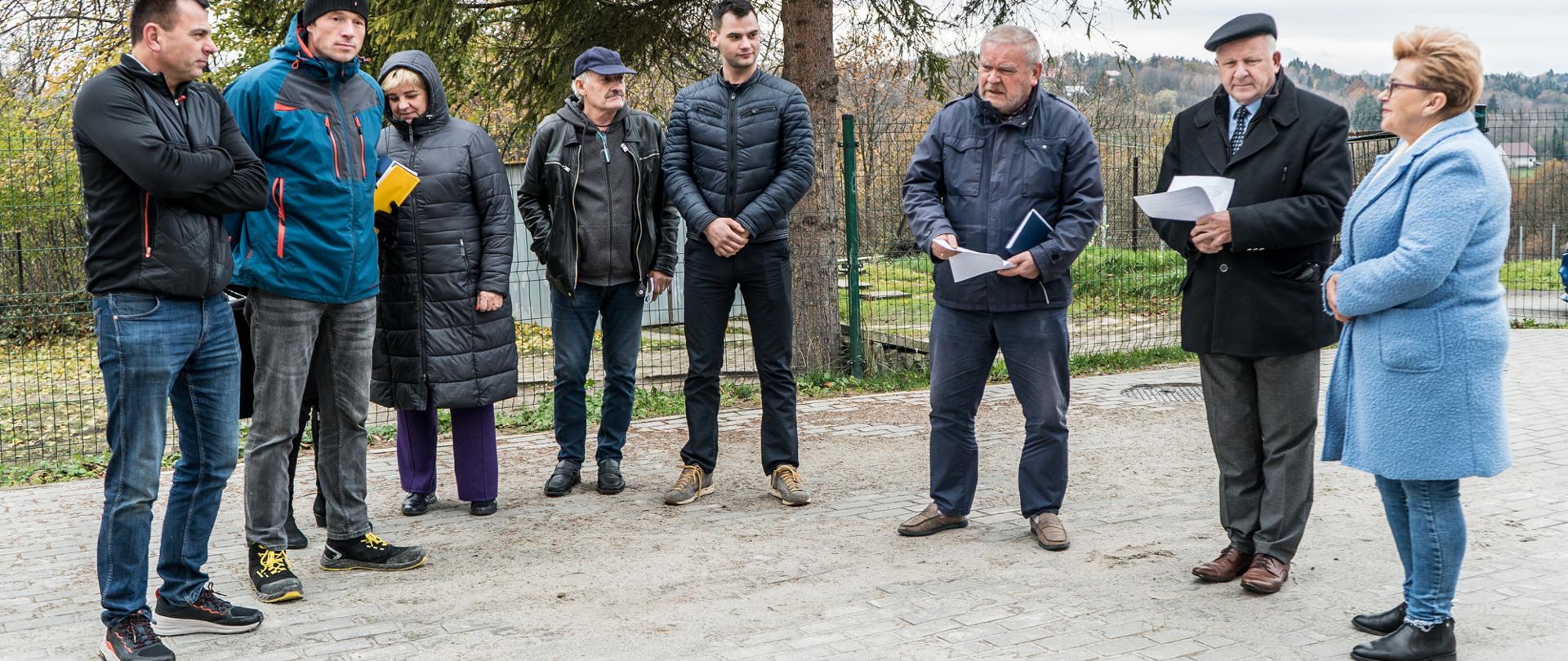 Na fotografii znajduje się osiem osób ustawionych w półokręgu, osoby te ubrane są w zimowe ubrania, toją na pustym placu i rozmawiają.