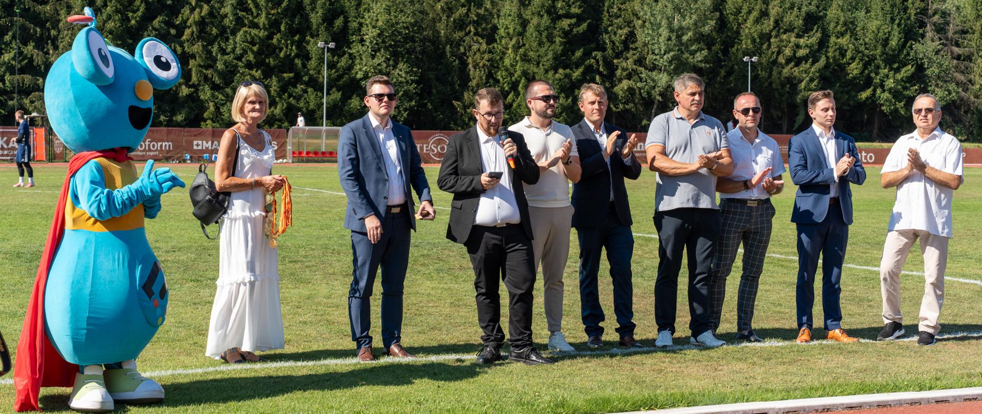 Piknik Kibica MKS Kalwarianka w Kalwarii Zebrzydowskiej – grupa uczestników i zaproszonych gości stojąca na murawie stadionu podczas uroczystości, obok klubowej maskotki. Jedna z osób trzyma medale przygotowane do wręczenia.