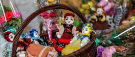 Kosz z lalkami w ludowych strojach otoczony kwiatami z bibuły i wielkanocnymi palmami.