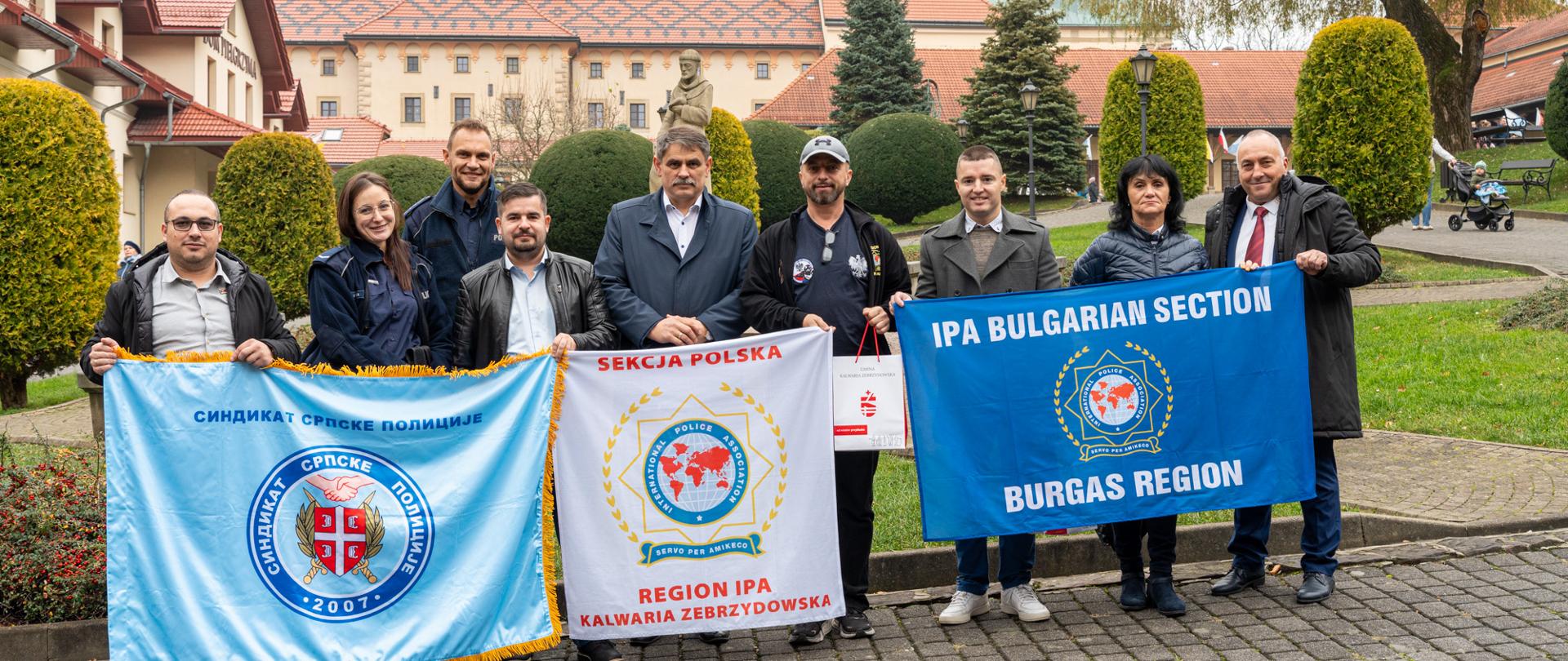 Na zdjęciu widać grupę przedstawicieli trzech sekcji IPA: Kalwaria Zebrzydowska (Polska), Burgas (Bułgaria) oraz Novi Sad / przedstawicieli serbskich służb, stojących wspólnie na tle Sanktuarium w Kalwarii Zebrzydowskiej. Uczestnicy trzymają trzy flagi swoich organizacji, prezentując współpracę i charakter delegacji. W centrum stoi Burmistrz Kalwarii Zebrzydowskiej Tadeusz Stela, otoczony przez polskich, bułgarskich i serbskich gości. Zdjęcie ma oficjalny, reprezentacyjny charakter – pokazuje międzynarodowe spotkanie, dobrą atmosferę i partnerskie relacje między delegacjami.
