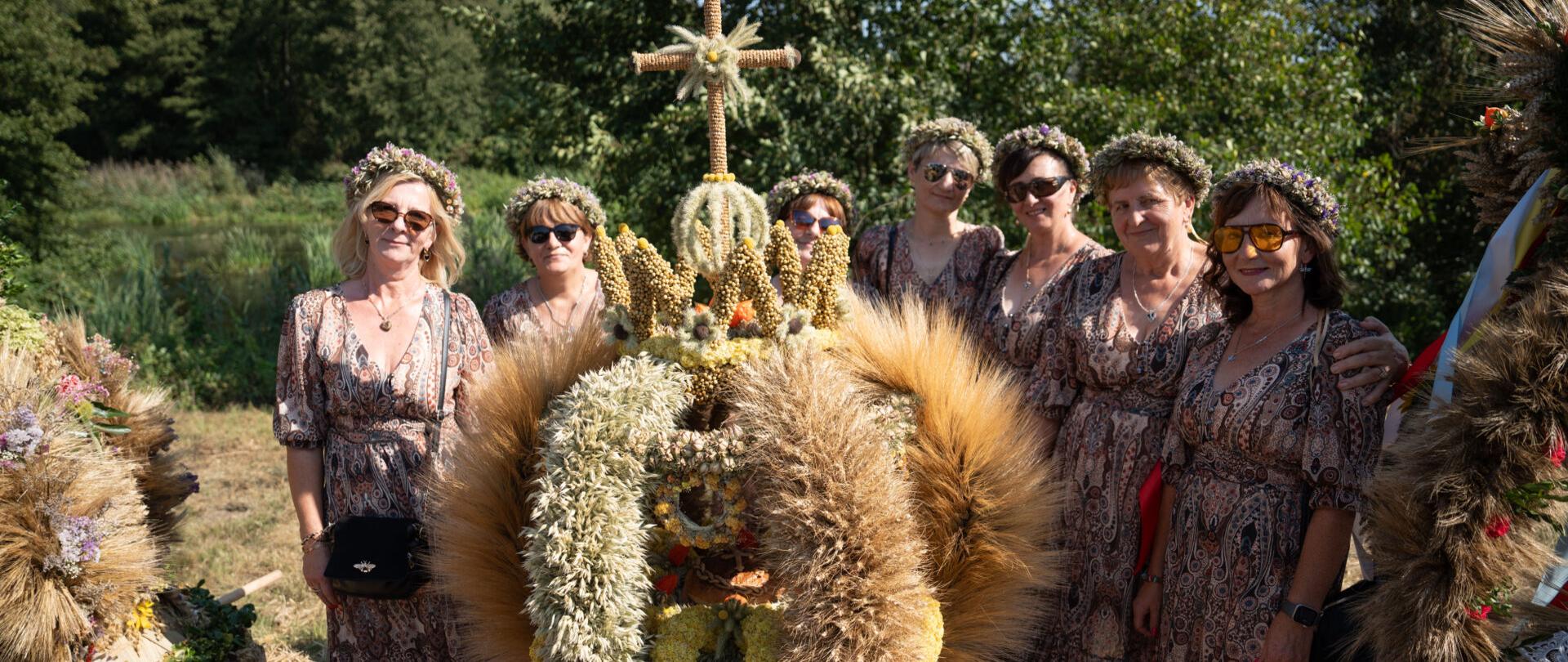 Siedem kobiet w dożynkowych strojach z wiankami z kwiatów na głowach. Przed nimi duży wieniec dożynkowy.