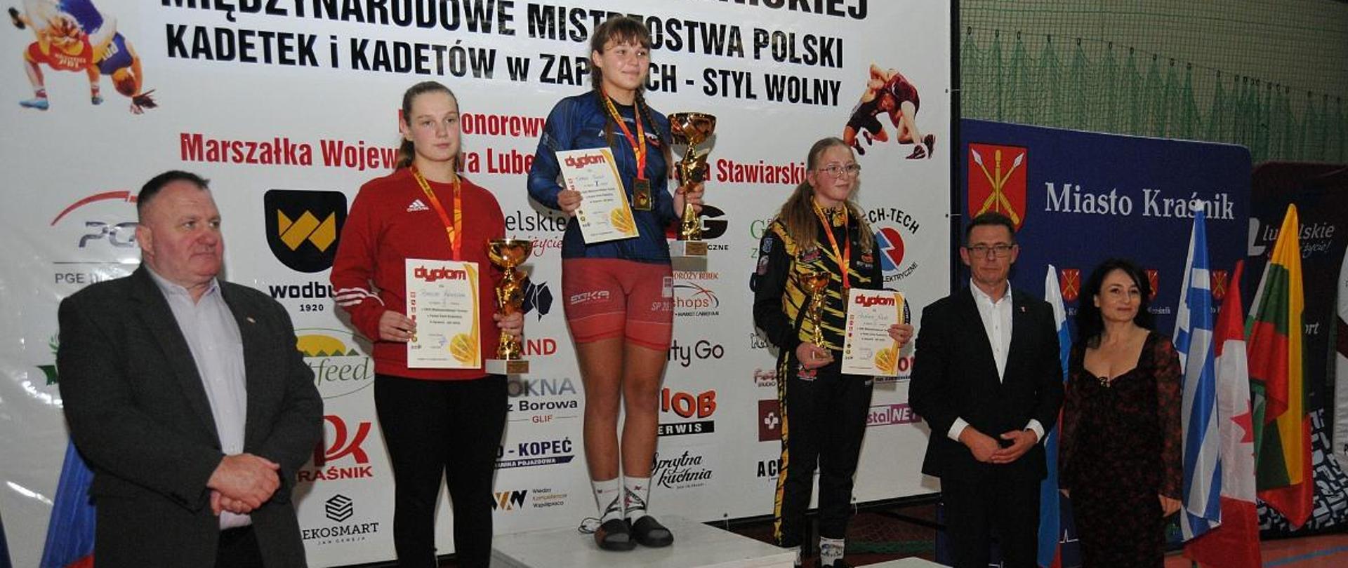 Trzy zawodniczki na podium po wręczeniu nagród w jednej z kategorii wagowych. Każda z nich ma zawieszony na szyi medal, a w rękach trzyma dyplom i puchar. Po lewej stronie podium stoi pan Jerzy Nieć, natomiast po prawej stronie zaraz przy trzecim stopniu podium stoi Przewodniczący Rady Powiatu w Kraśniku Jacek Dubiel.
Zdjęcie: Krasnik24.pl