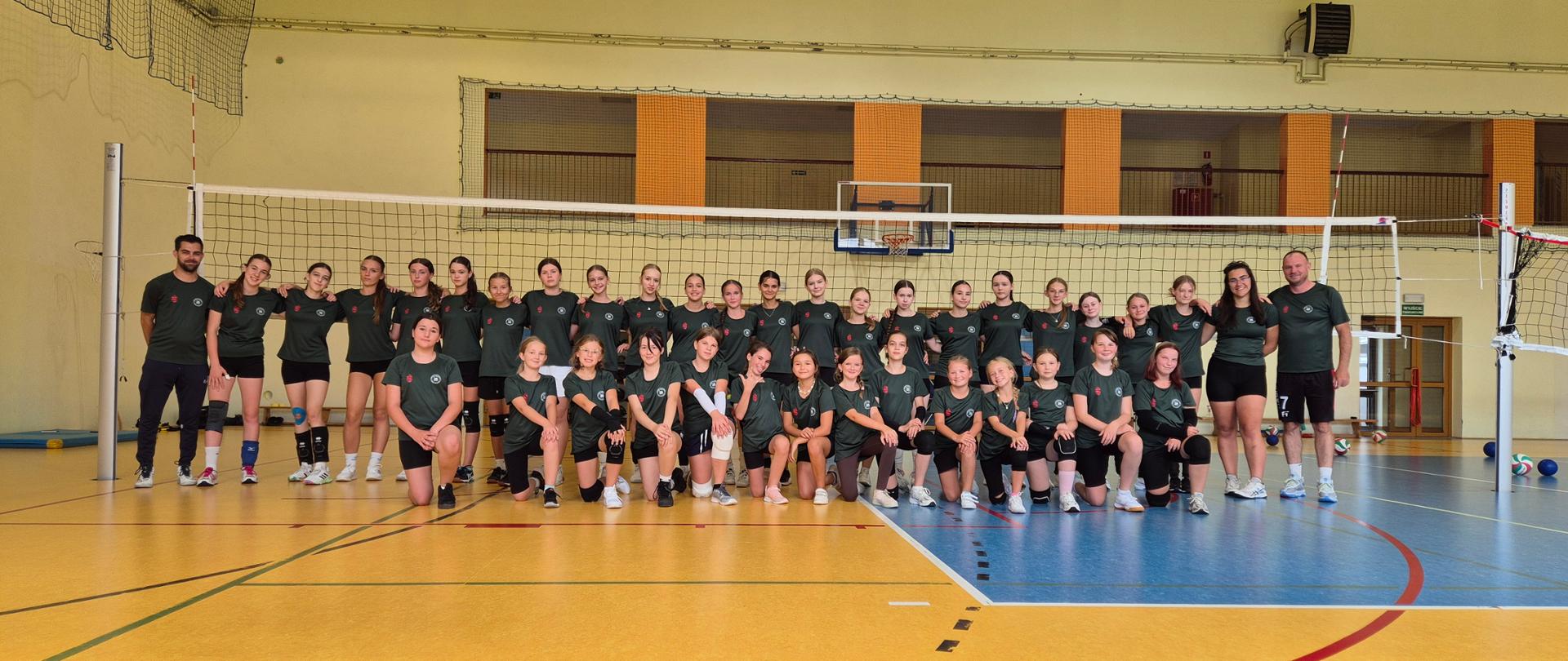 Na zdjęciu widać dużą grupę dziewcząt i kobiet w zielonych koszulkach sportowych, ustawionych w dwóch rzędach na hali sportowej. Za nimi znajduje się siatka do siatkówki, co sugeruje, że jest to drużyna lub grupa treningowa siatkarek. W grupie znajdują się także dwaj trenerzy (po lewej i prawej stronie). Tło stanowi ściana hali z koszem do koszykówki i balkonem dla widzów. Całość wygląda jak zdjęcie pamiątkowe wykonane po treningu lub turnieju.