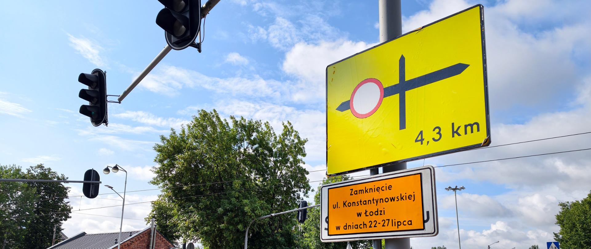 Skrzyżowanie z sygnalizacją świetlną oraz żółtym znakiem informującym o zamknięciu drogi za 4,3 km. Poniżej znajduje się tablica z informacją o zamknięciu ulicy Konstantynowskiej w Łodzi w dniach 22-27 lipca. W tle widoczne są drzewa, niska zabudowa miejsca, kolejne sygnalizatory oraz częściowo zachmurzone niebo.
