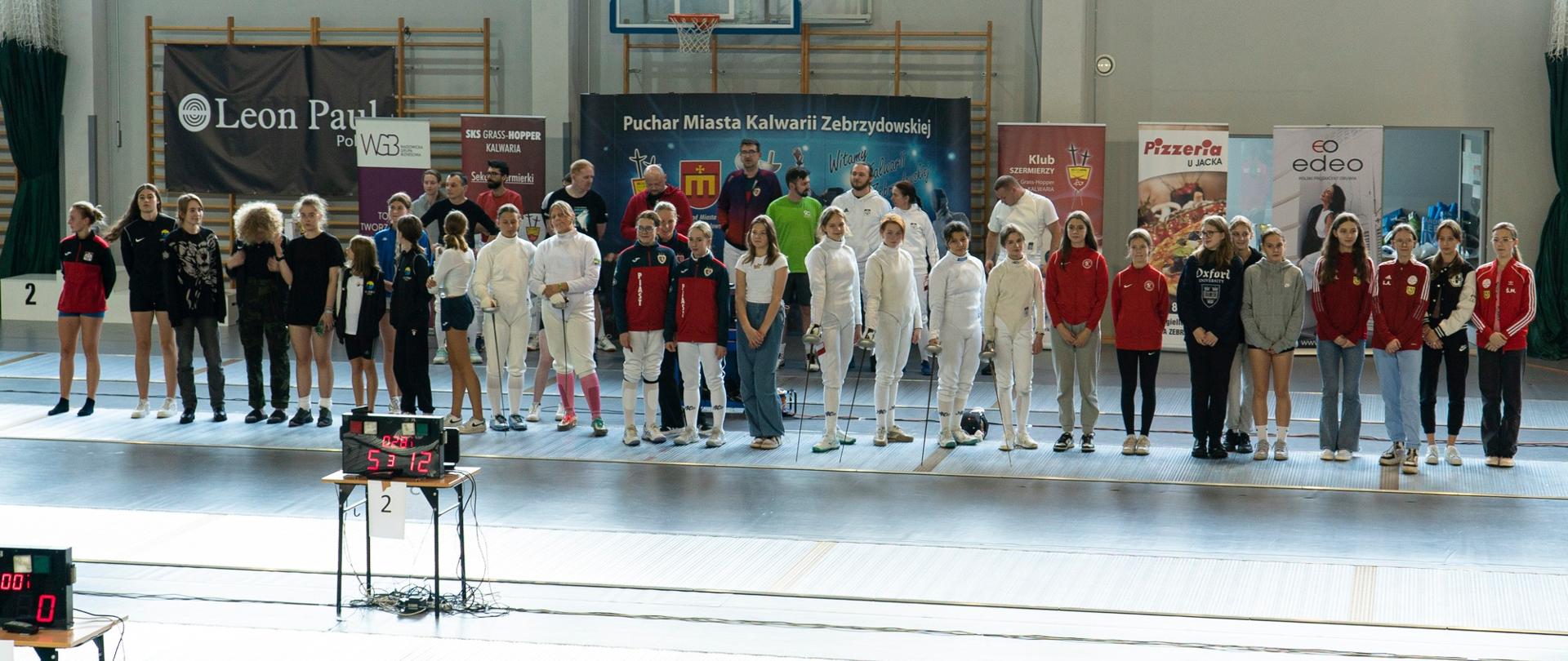 W hali sportowej do wspólnego zdjęcia pozują zawodnicy turnieju i ich trenerzy; przed nimi elektroniczna plansza do wyświetlania punktacji; w tle banery i rollupy organizatorów i sponsorów