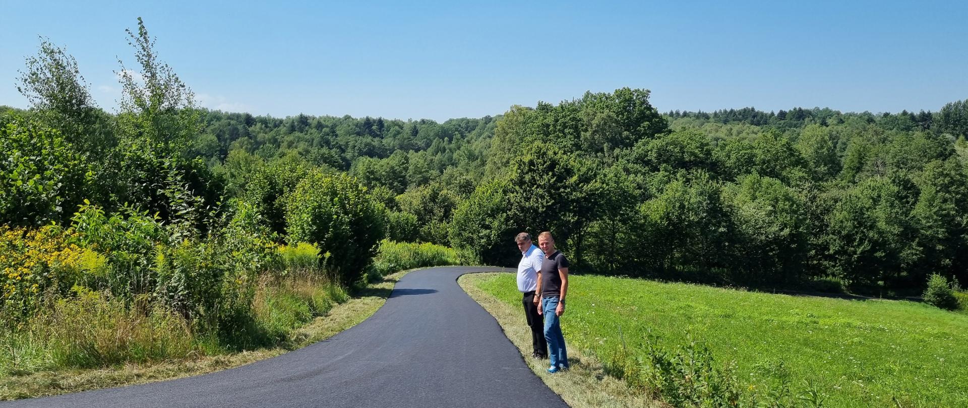 Na zdjęciu widać świeżo wyasfaltowaną, wąską drogę wijącą się przez zielone tereny. Po prawej stronie drogi stoją dwaj mężczyźni – jeden w białej koszuli, drugi w czarnej koszulce – patrzący w stronę aparatu. W tle rozciąga się panorama gęstego lasu pod błękitnym, bezchmurnym niebem. 