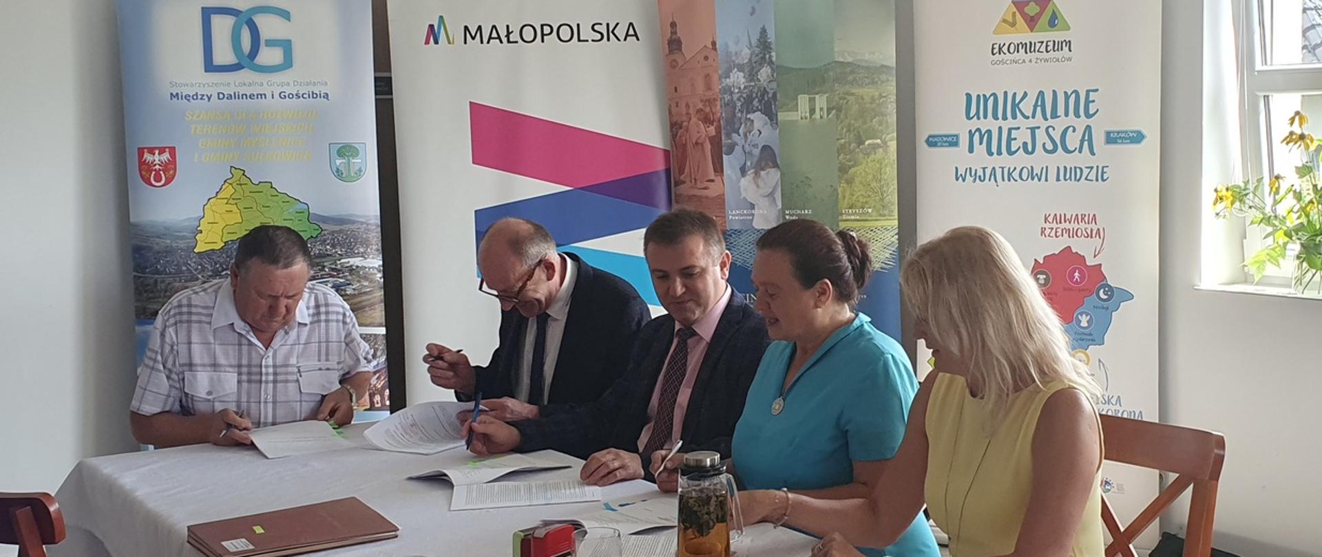 Przy stole z dokumentami siedzą przedstawiciele stron podpisujących umowę, za nimi banery lokalnych grup działania i województwa
