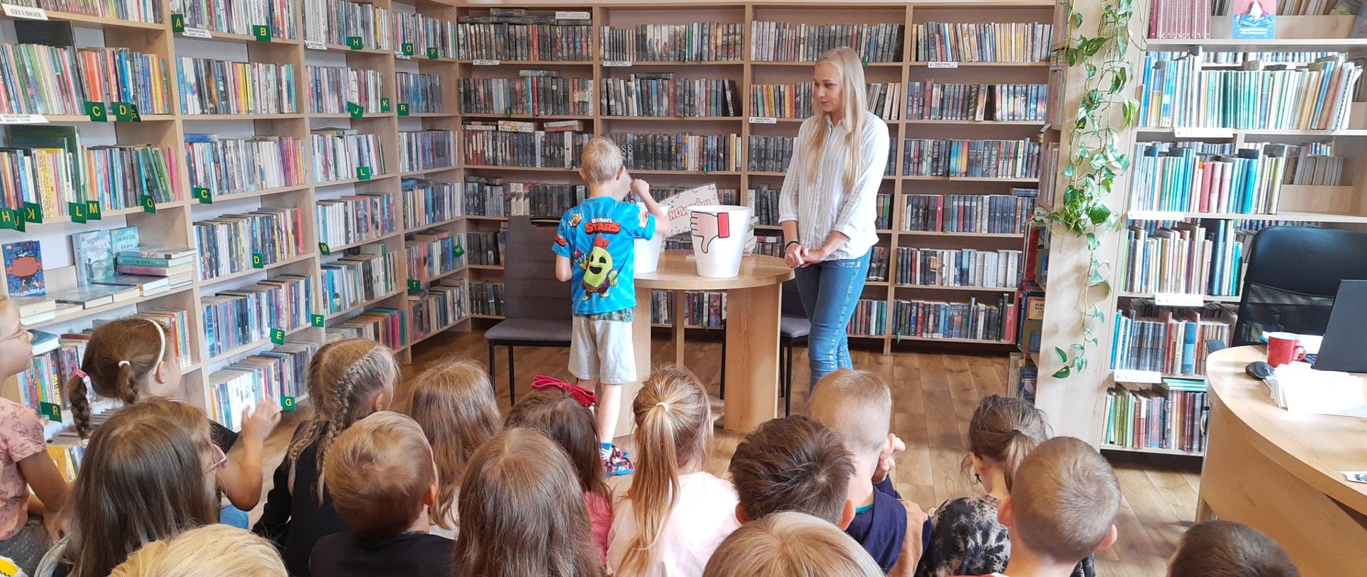 Dzieci w bibliotece uczestniczą w zajęciach. W tle regały z książkami.