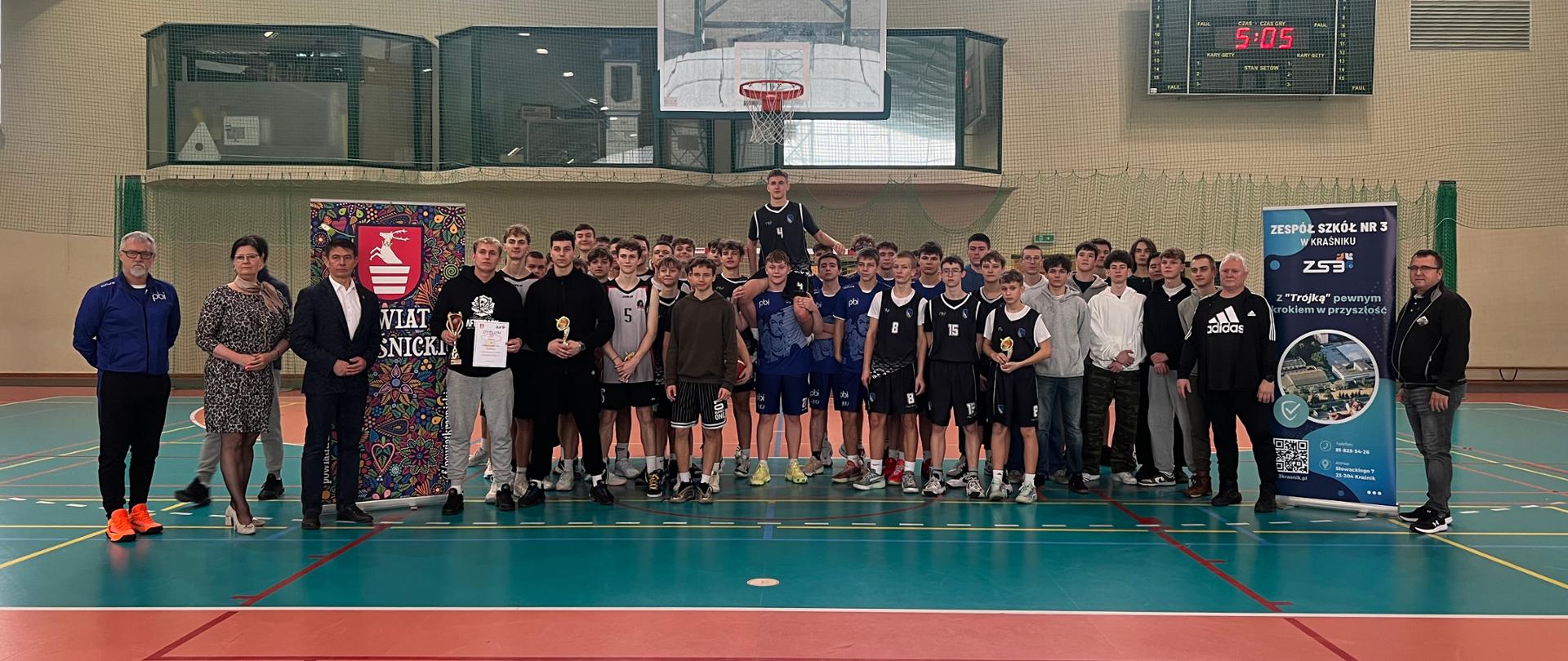 Pośrodku hali sportowej stoją zawodnicy biorący udział w turnieju, ich opiekunowie i trenerzy, Przewodniczący Rady Powiatu w Kraśniku Jacek Dubiel i Dyrektor Zespołu Szkół nr 3 w Kraśniku Anna Gromek. Po lewej stronie ustawiony jest rollaup Starostwa Powiatowego w Kraśniku, a po stronie prawej - rollup Zespołu Szkół nr 3 w Kraśniku.