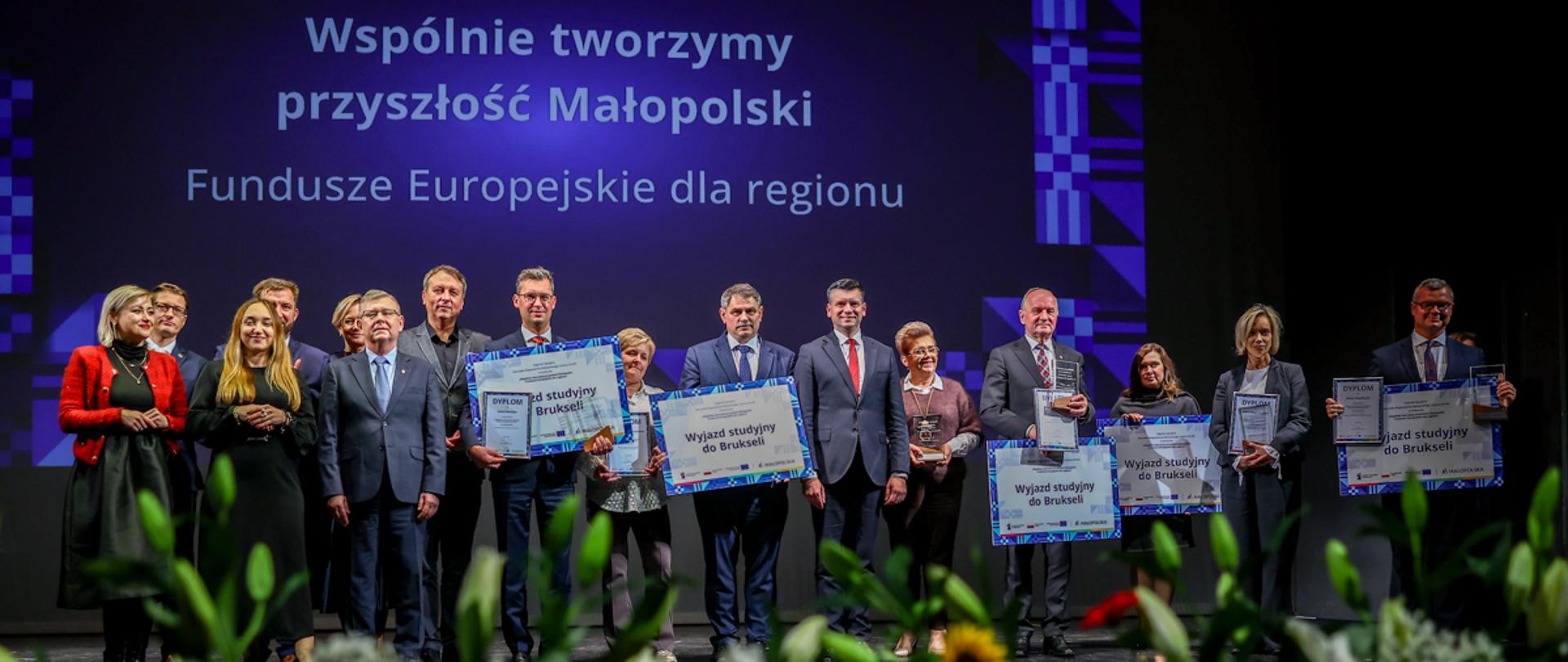 Uroczysta gala wręczenia nagród w konkursie 'Wspólnie tworzymy przyszłość Małopolski – Fundusze Europejskie dla regionu'. Na scenie stoi grupa laureatów i organizatorów z dyplomami oraz tablicami z napisem 'Wyjazd studyjny do Brukseli'. W tle widoczny jest ekran z nazwą wydarzenia, a na pierwszym planie kompozycja z kwiatów – słoneczników, róż i lilii. 