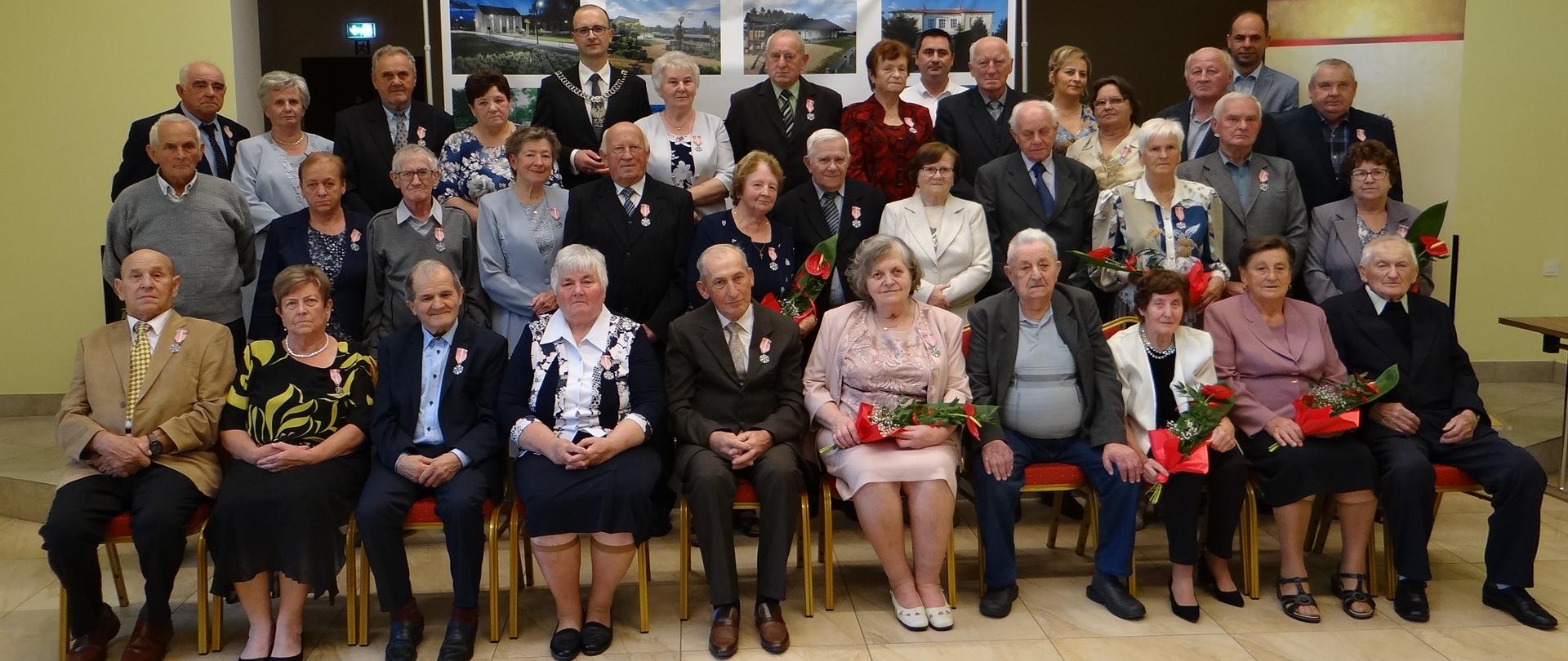 Dostojni Jubilaci 50 i 60 lat pożycia małżeńskiego. Zbiorowa fotografia wykonana w sali widowiskowej Gminnej Biblioteki Publicznej. Osoby w pierwszym rzędzie siedzą na krzesłach, dwa dalsze rzędy stoją. 