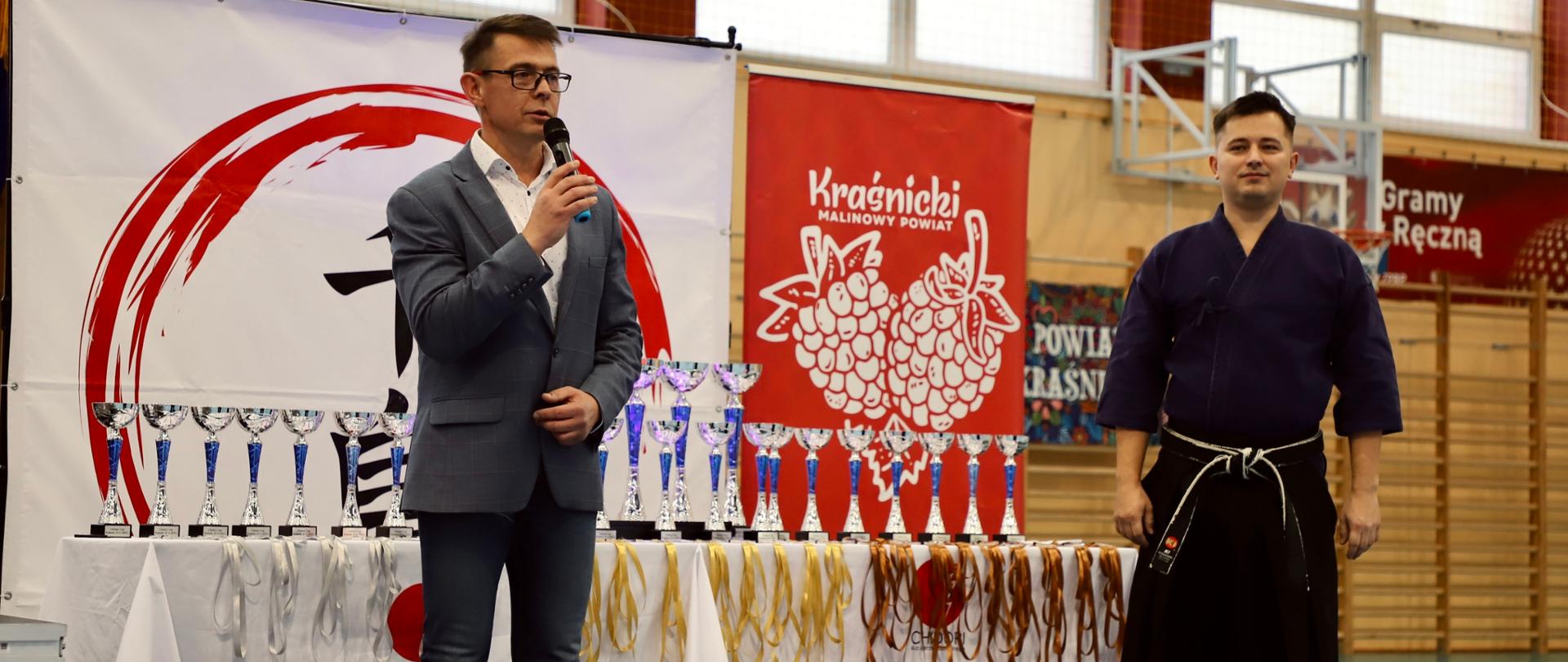 Przewodniczący Rady Powiatu w Kraśniku Jacek Dubiel podczas przemówienia (pierwszy z lewej). Obok stoi trener i jednocześnie prezes Kraśnickiego Klubu Karate Tradycyjnego Chidori w Kraśniku Marek Bury. Za nimi ustawiony jest stolik, na którym znajdują się medale i puchary.