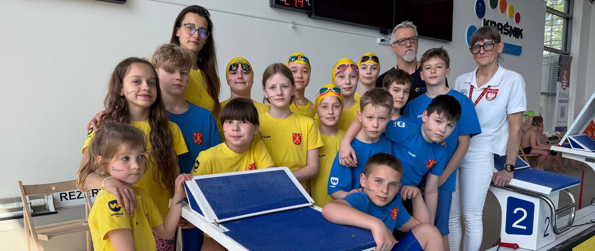 15-osobowa grupa zawodników Uczniowskiego Klubu Pływackiego "Fala" w Kraśniku w strojach żółtych i niebieskich stoi przy ścianie pływalni wraz z trenerami: Zuzanną Mazurek, Iwoną Mazurek i Grzegorzem Mazurkiem.