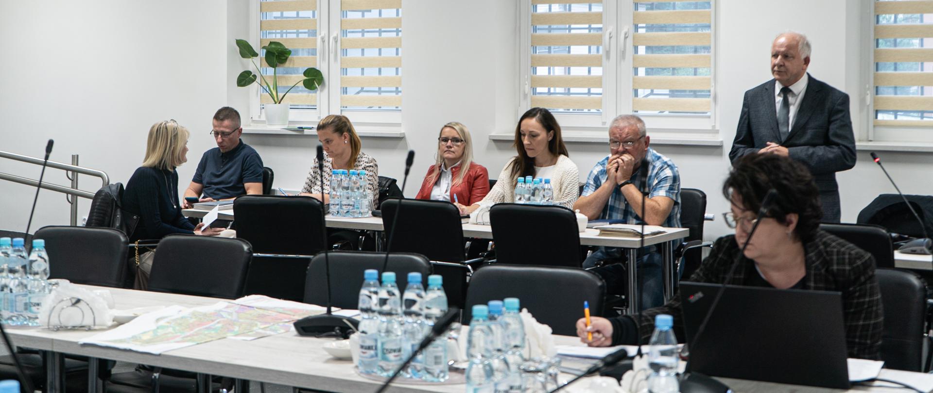 W jasnej sali posiedzeń przy stolikach siedzą pracownicy urzędu; na stołach znajdują się mikrofony i woda w butelkach; po prawej stronie kadru burmistrz przemawia stojąc.