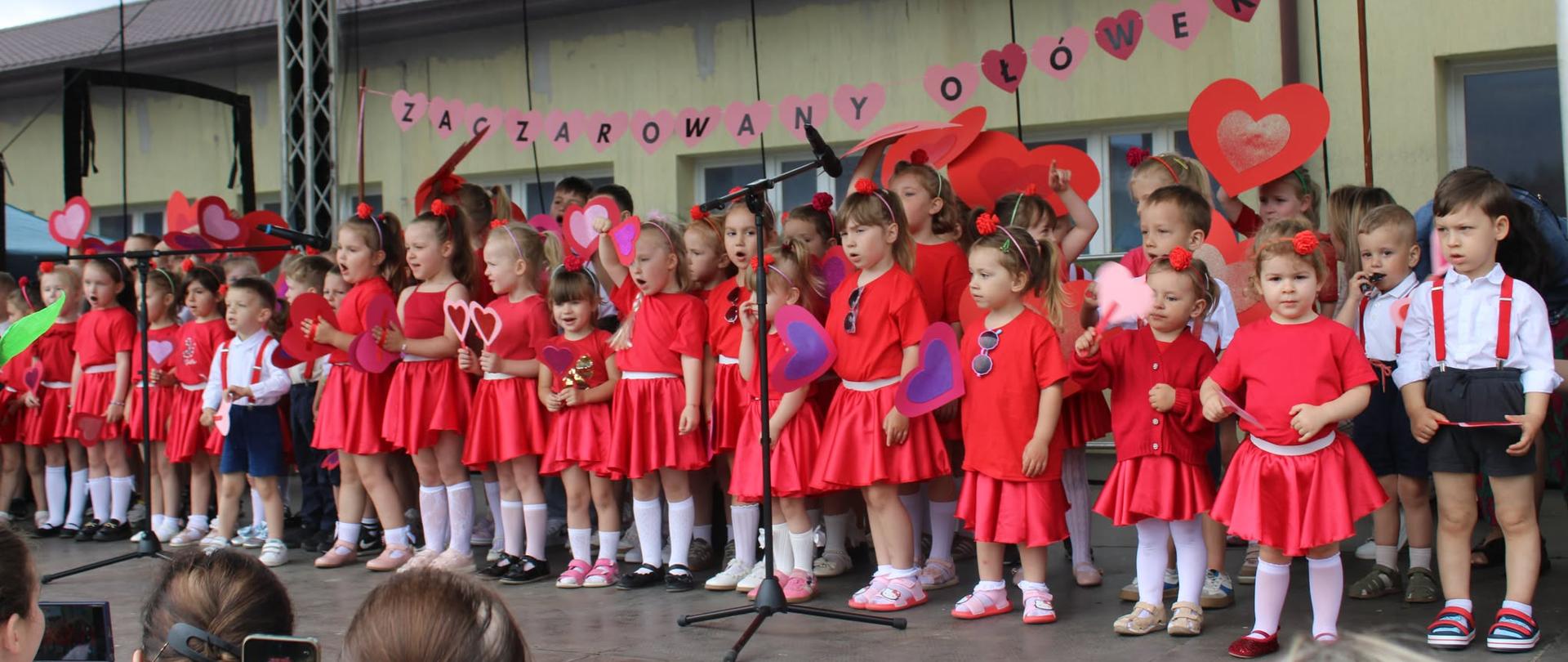 Duża grupa małych dzieci (około 40) w czerwonych sukienkach i białych strojach z czerwonymi szelkami wykonuje spektakl na scenie plenerowej. Dzieci trzymają kolorowe serca z papieru. W tle baner z napisem "Zaczarowany Ołówek". Publiczność fotografuje wydarzenie.