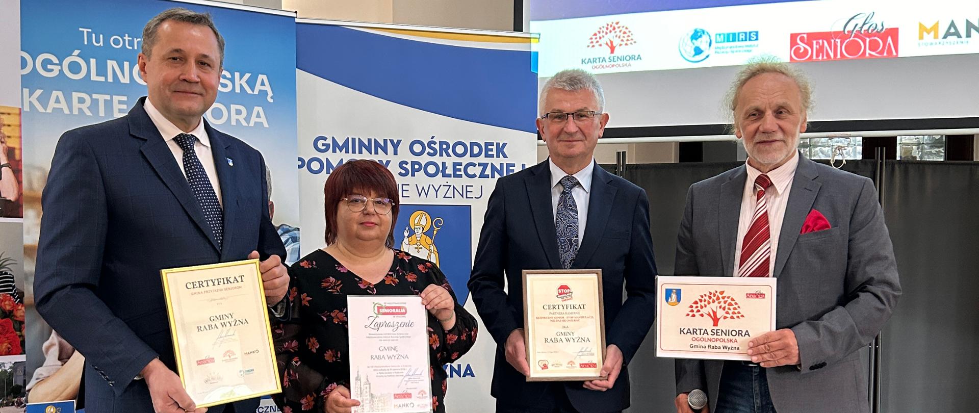 Przedstawiciele władz gminy pozuje do zdjęcia, trzymając certyfikat „Gmina Przyjazna Seniorom”. W tle widoczne są banery, prezentacja multimedialna oraz materiały promujące inicjatywę.