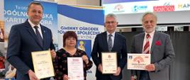 Przedstawiciele władz gminy pozuje do zdjęcia, trzymając certyfikat „Gmina Przyjazna Seniorom”. W tle widoczne są banery, prezentacja multimedialna oraz materiały promujące inicjatywę.