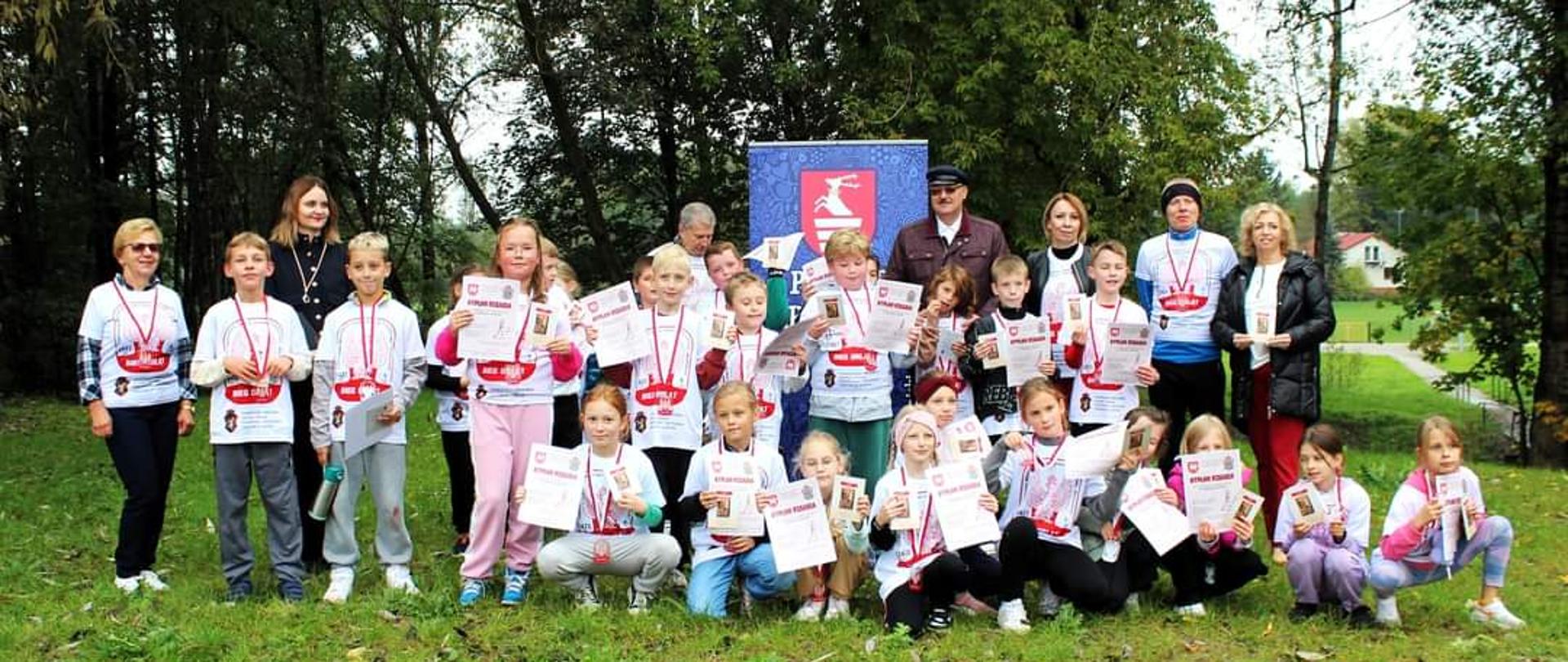 Uczestnicy biegu, stojący na boisku Gminnego Klubu Sportowego "Orzeł" Urzędów. Z tyłu za nimi stoi rollup Starostwa Powiatowego w Kraśniku, a na dalszym planie widać drzewa. Zawodnicy mają założone białe okolicznościowe koszulki, na szyjach zawieszone medale, a w rękach trzymają dyplomy.