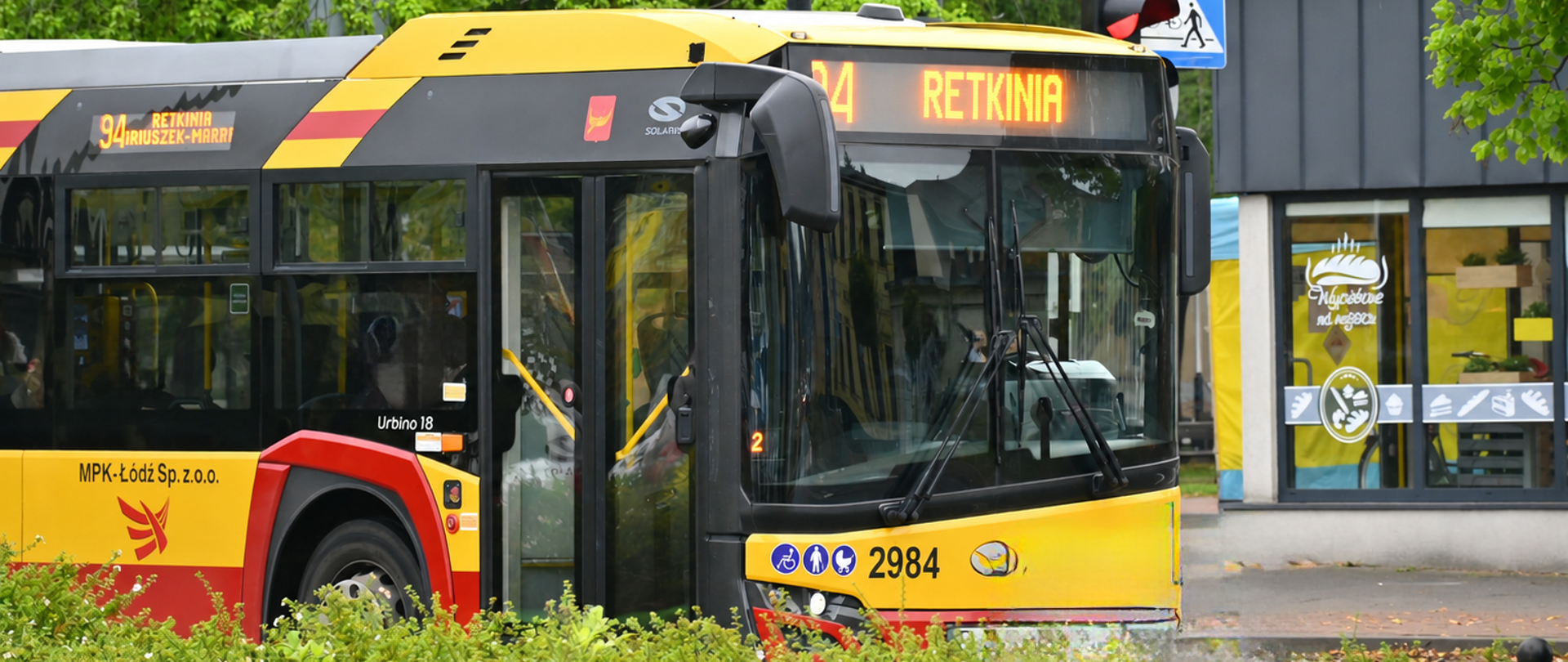 Żółto-czerwony autobus linii 94 jadący w kierunku Retkini stoi przy skrzyżowaniu w otoczeniu zielonych drzew i znaków drogowych.