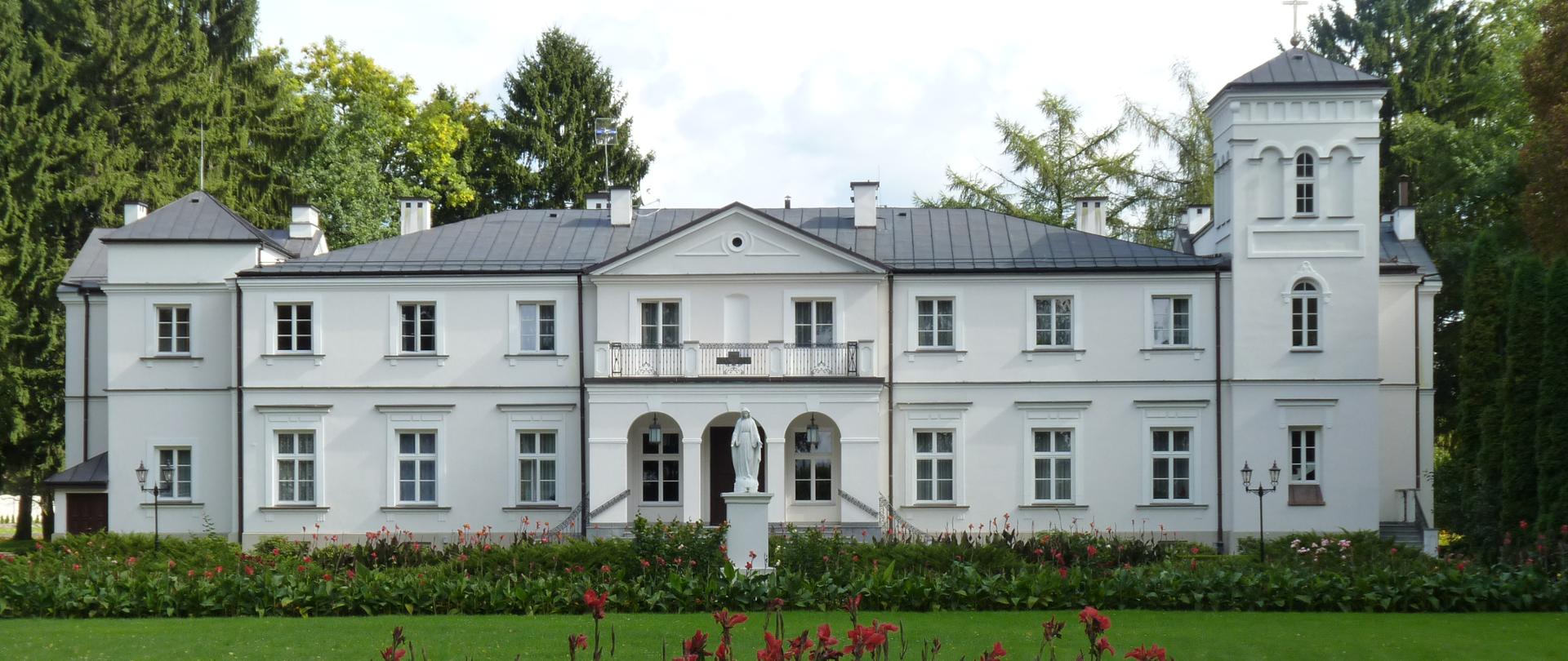 Duży biały budynek. Przed budynkiem stoi biała figura w kształcie człowieka