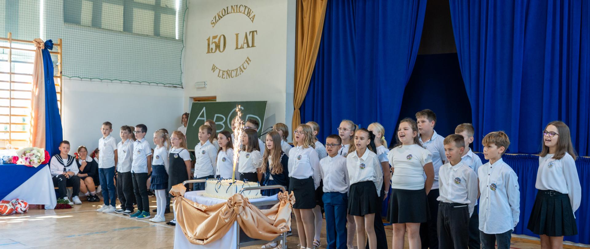 Uczniowie Zespołu Szkół nr 6 w Leńczach występują podczas uroczystości jubileuszu 150-lecia szkolnictwa w Leńczach. Dzieci stoją przed sceną w szkolnej sali gimnastycznej, a przed nimi znajduje się jubileuszowy tort z zapaloną fontanną. W tle widoczny jest napis „150 lat szkolnictwa w Leńczach” oraz niebiesko-złota dekoracja sceny.