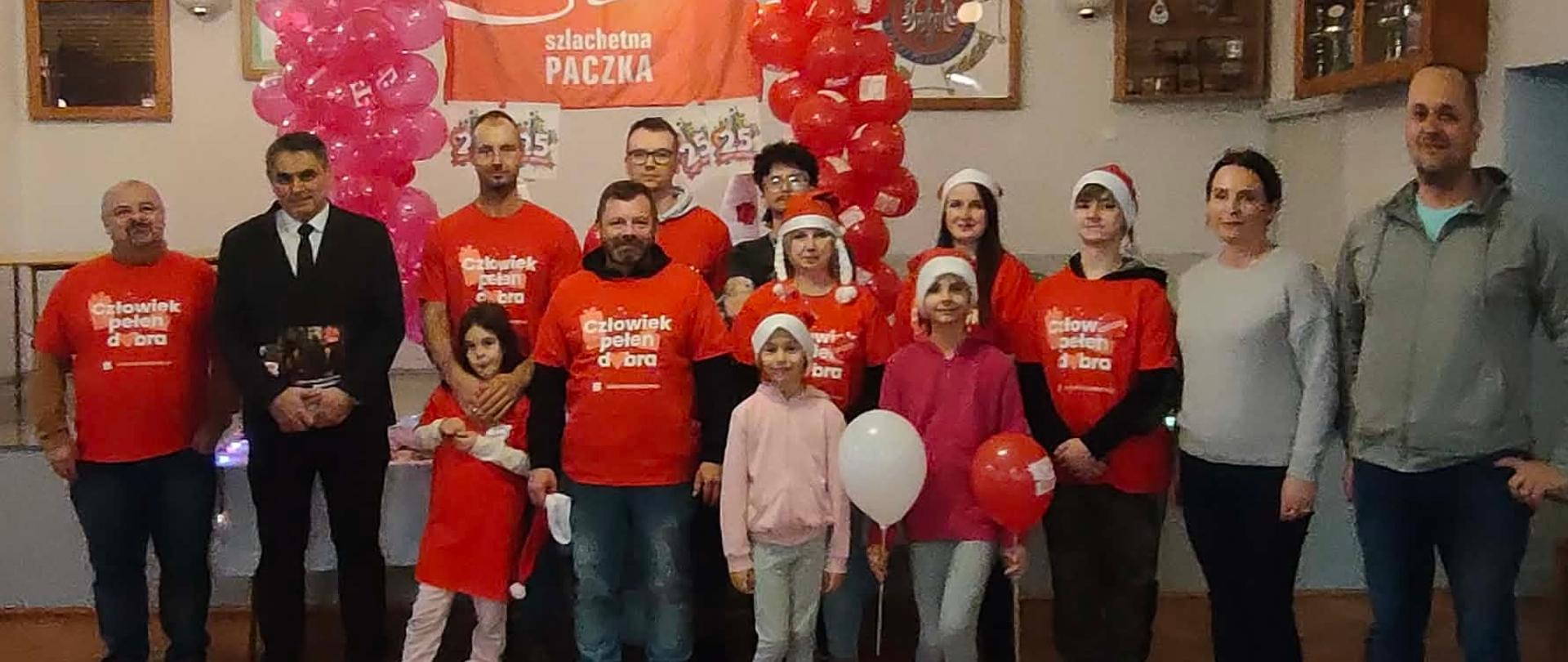 Grupowe zdjęcie uczestników akcji Szlachetna Paczka wykonane w sali świetlicowej. Kilkanaście osób dorosłych i dzieci stoi w jednym rzędzie. Większość dorosłych ma na sobie czerwone koszulki z napisem „Człowiek pełen dobra”, część dzieci trzyma czerwone i białe balony, niektórzy mają czapki świąteczne. W tle widoczny jest pomarańczowy baner „Szlachetna Paczka” oraz dwie kolumny balonów w kolorze różowym i czerwonym. Zdjęcie ma świąteczny i wspólnotowy charakter.