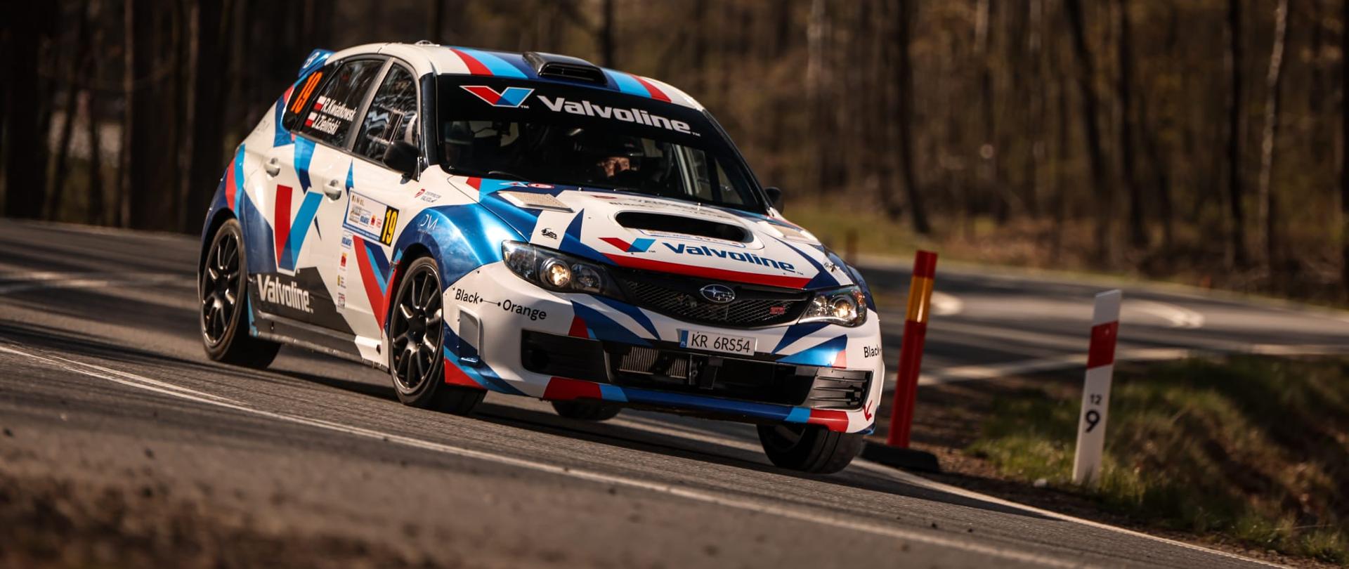 Samochód w kolorach biało czerwono niebieskich mknie na zakręcie, jeden z etapów rajdu.