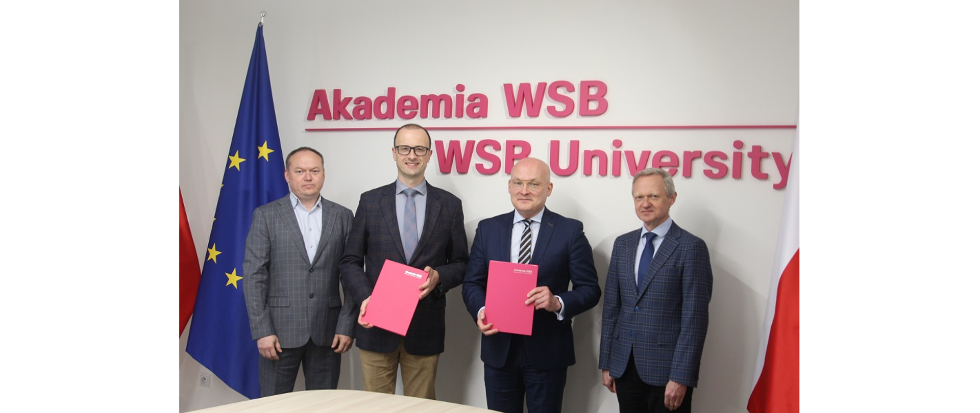 Sygnatariusze porozumienia pomiędzy Gminą Hażlach a Akademią WSB w Dąbrowie Górniczej. Fot. Akademia WSB. Stoją czterej mężczyźni, dwaj z nich trzymają teczki z podpisanymi umowami. Obok flagi UE i RP. Z tyłu na ścianie napis Akademia WSB, WSB University. 