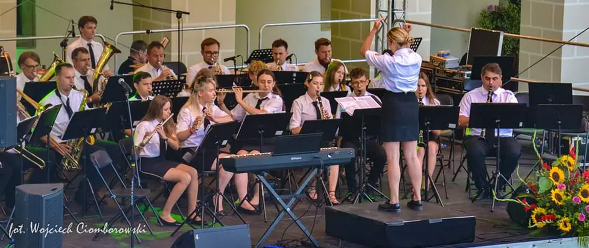 Orkiestra dęta na pierwszym planie dyrygent, siedzą członkowie orkiestry w białych koszulach z instrumentami. 