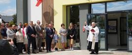 Poświęcenie nowej biblioteki przez księdza proboszcza Jana Balickiego. Duchowny stoi przed wejściem, unosi rękę z kropidłem. Obok niego zgromadzeni goście, w tym przedstawiciele władz i mieszkańcy Giedlarowej.