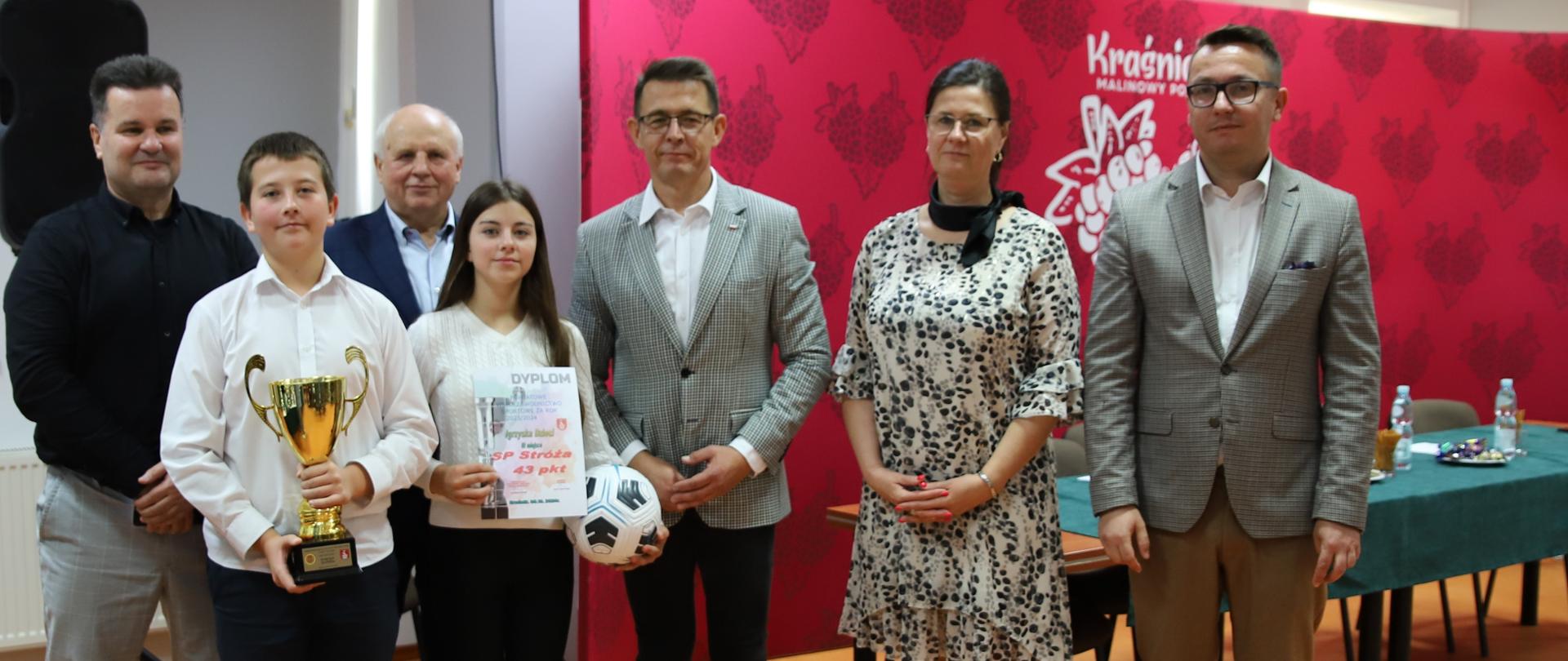 Dwóch uczniów Szkoły Podstawowej w Stróży wraz z nauczycielem wychowania fizycznego (od lewej) trzymająca w rękach puchar, dyplom i piłkę. Od prawej strony stoją: Sekretarz Miasta Kraśnik Łukasz Skokowski, Dyrektor Zespołu Szkół nr 3 w Kraśniku Anna Gromek, Przewodniczący Rady Powiatu w Kraśniku Jacek Dubiel i Przewodniczący Powiatowego Szkolnego Związku Sportowego w Kraśniku Zbigniew Gawdzik.