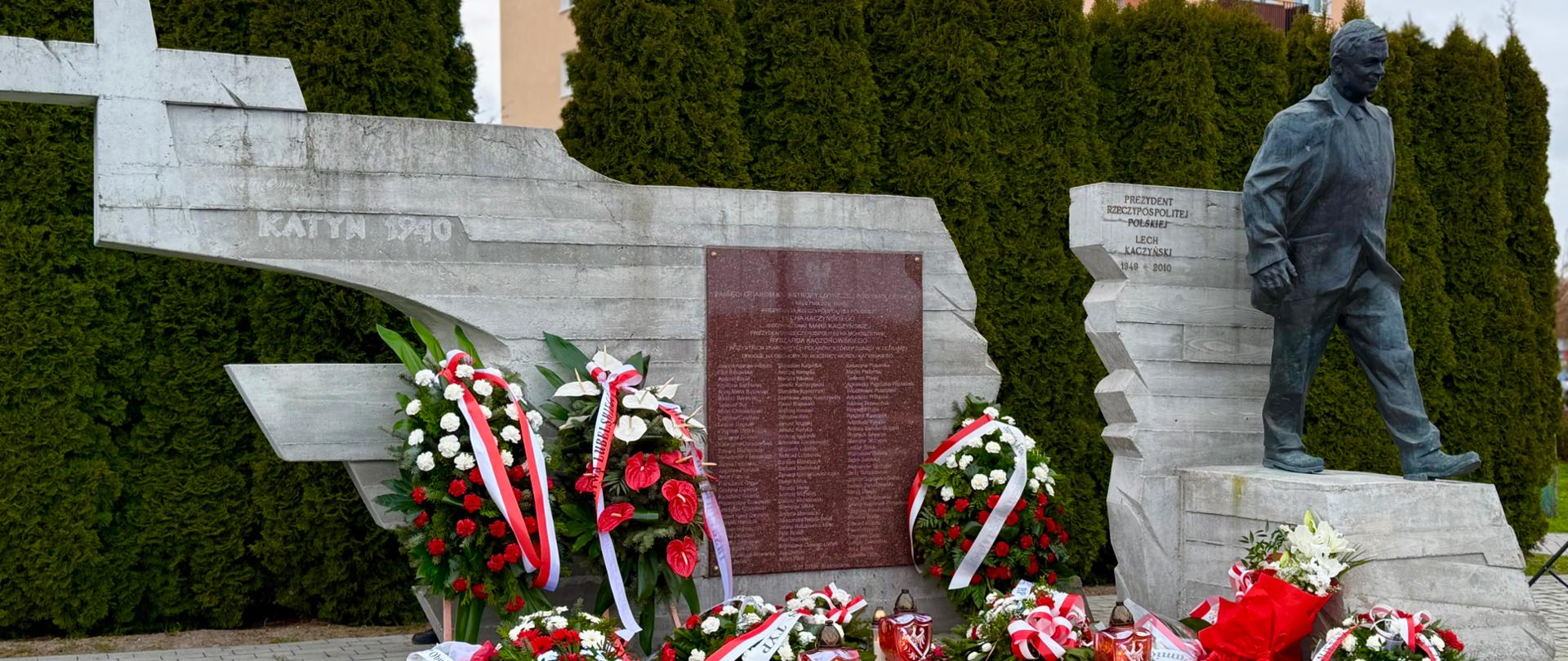 Pomnik upamiętniający ofiary katastrofy smoleńskiej i zbrodni katyńskiej – betonowa konstrukcja z sylwetką samolotu, listą ofiar oraz rzeźbą prezydenta Lecha Kaczyńskiego. Przed pomnikiem liczne wieńce, wiązanki i znicze ułożone w kształt krzyża. W tle widoczne są tuje oraz bloki mieszkalne.