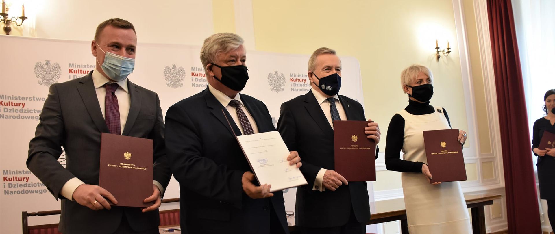 Troje mężczyzn w garniturach oraz kobieta trzymają w rękach przed sobą teczki z orłem