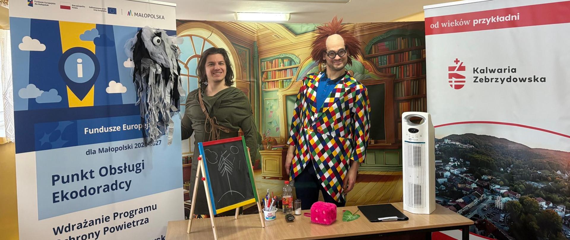 Dwaj aktorzy w kostiumach, przed nimi stolik z rekwizytami, po bokach banery (rollupy) promocyjne