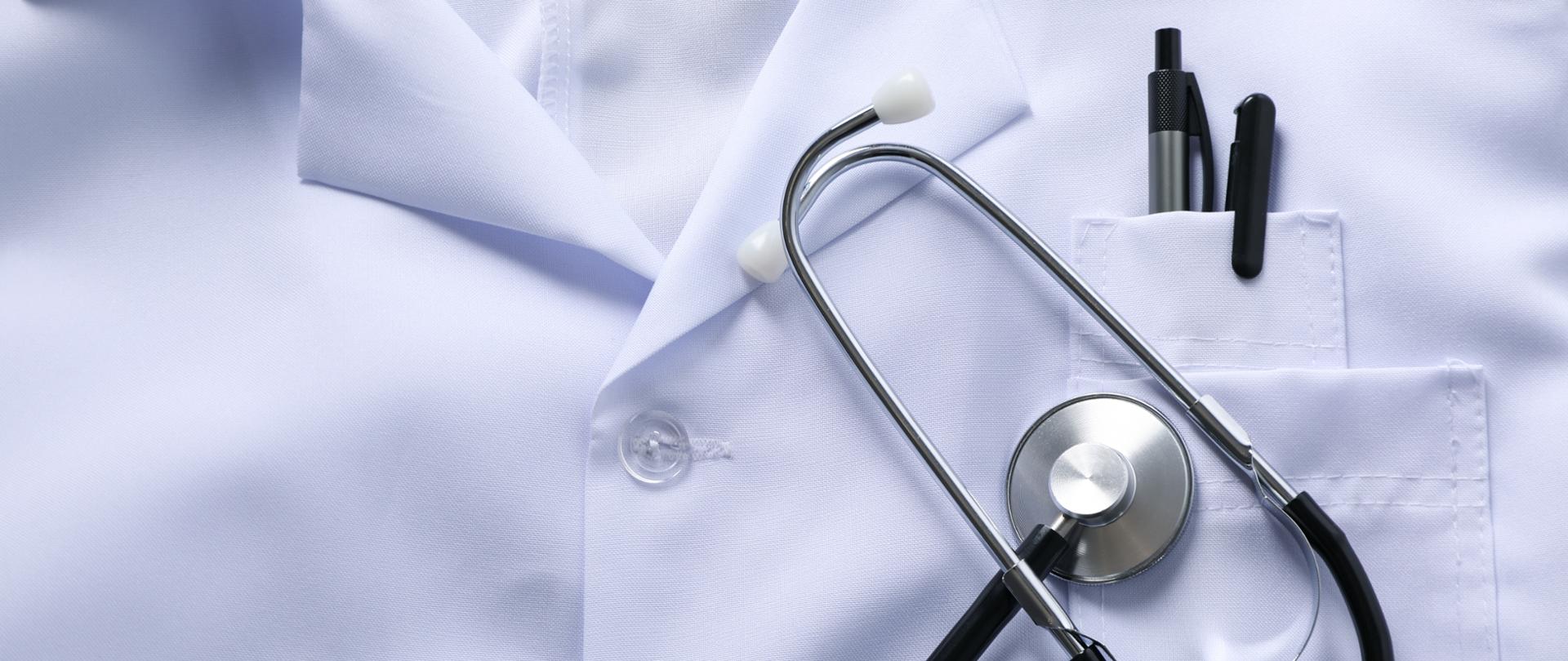 Zbliżenie na biały fartuch medyczny z kieszenią, w której znajdują się długopisy. Na fartuchu leży stetoskop, symbolizujący pracę lekarzy i personelu medycznego oraz opiekę zdrowotną.